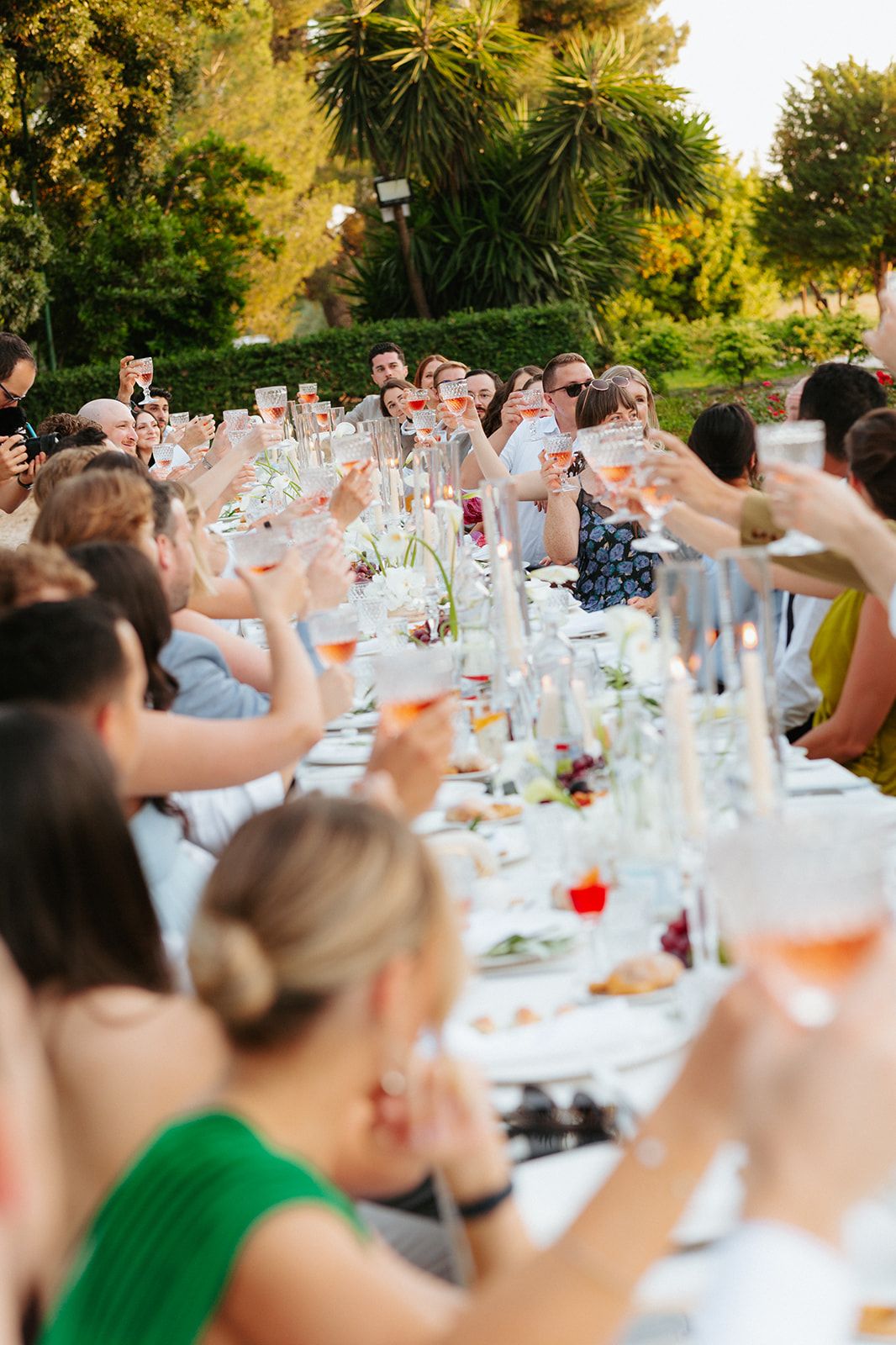 Wedding guests raise glasses in a toast during an outdoor garden reception at a long dining table with floral centerpieces.