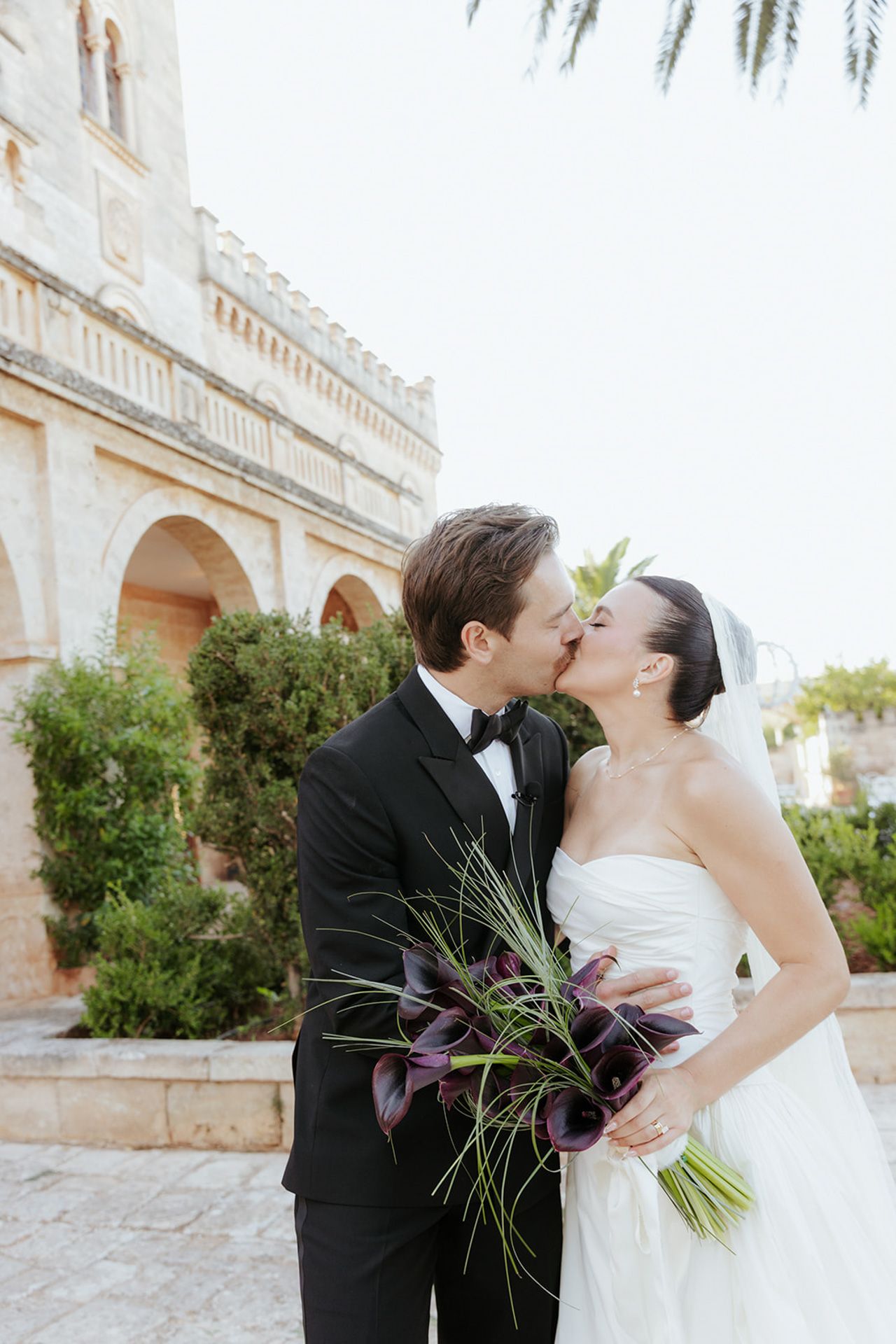 A couple shares a romantic kiss in front of a Mediterranean-style building with palm trees and elegant archways.