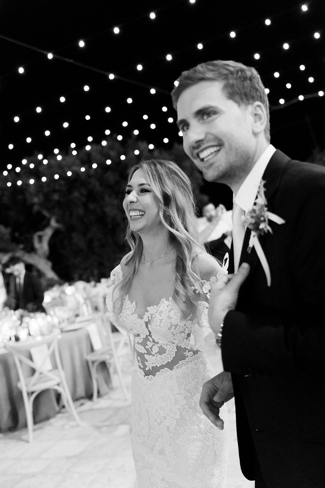 A joyful black and white wedding photo of newlyweds laughing together under string lights at their outdoor reception.