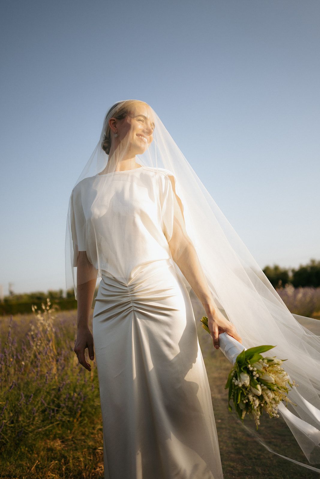 A serene bride in a simple white dress stands in a golden field at sunset as her veil flows gracefully in the breeze.