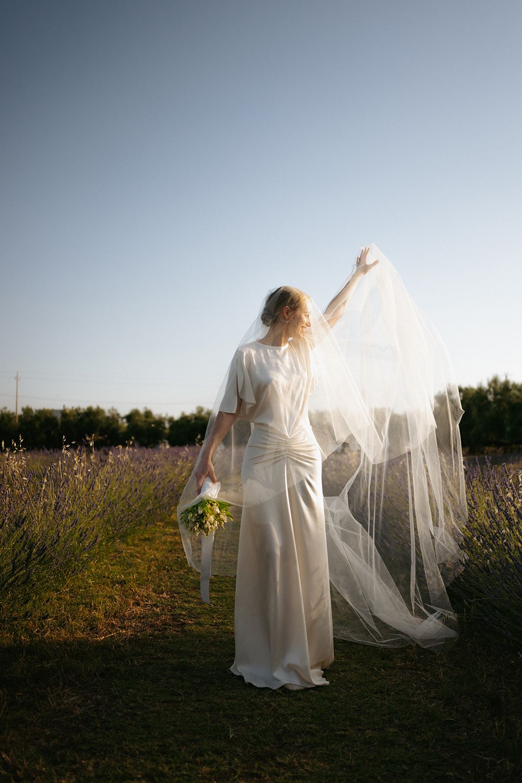 A person in a flowing white wedding dress holds a delicate veil while standing in a sunlit field with flowers.