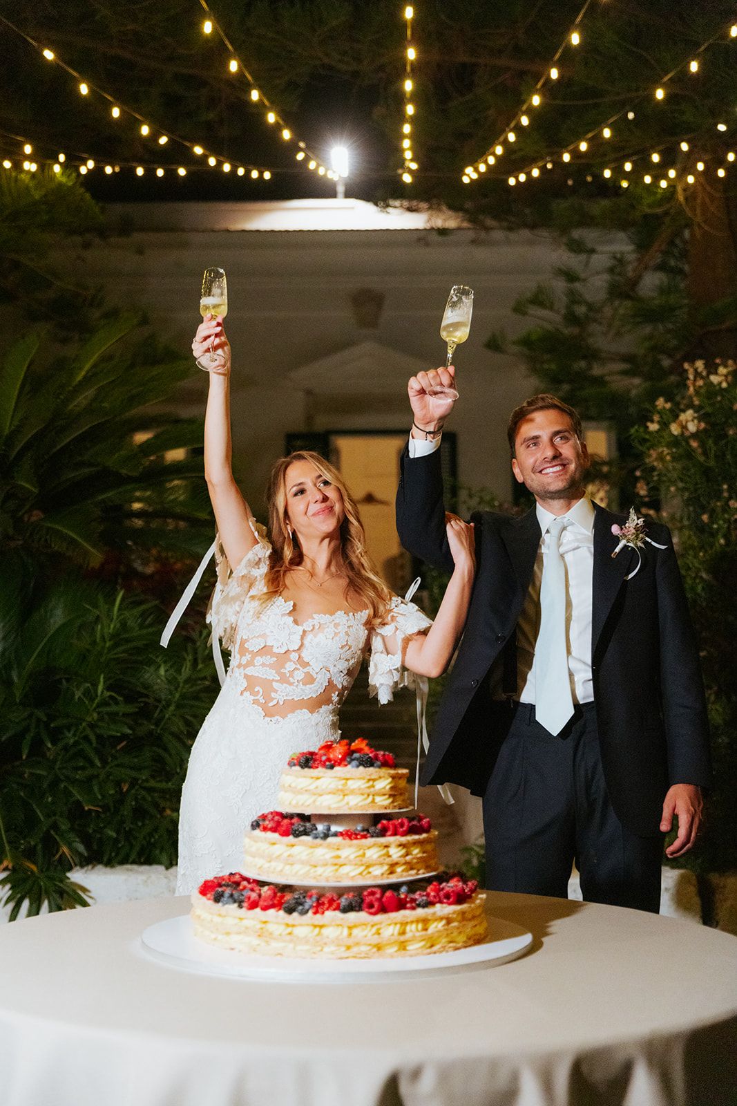 Couple celebrating at wedding reception with three tiered cake decorated with fresh berries under string lights.