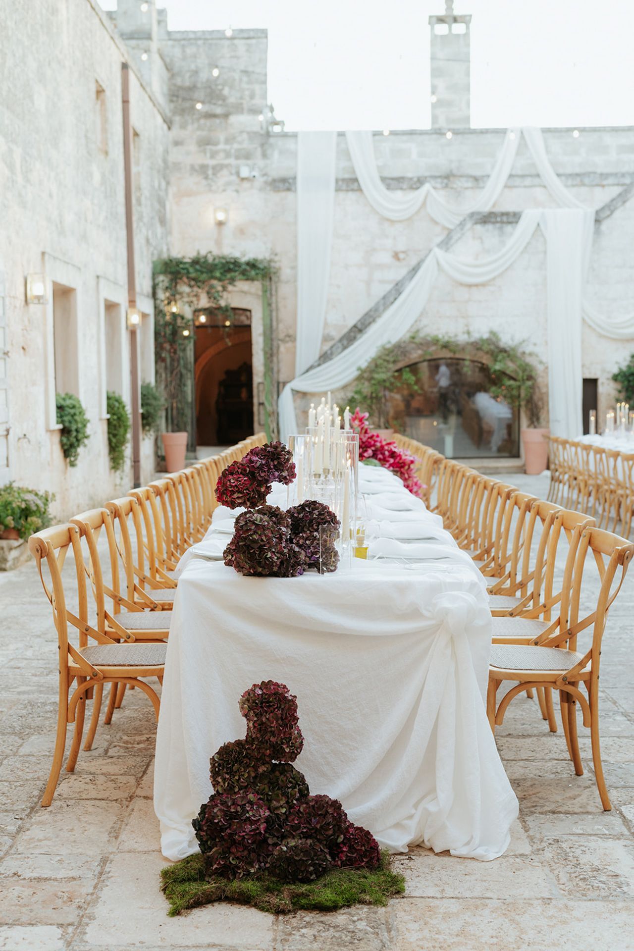 An elegant wedding reception setup with long tables, white linens and dark floral arrangements in a stone courtyard.