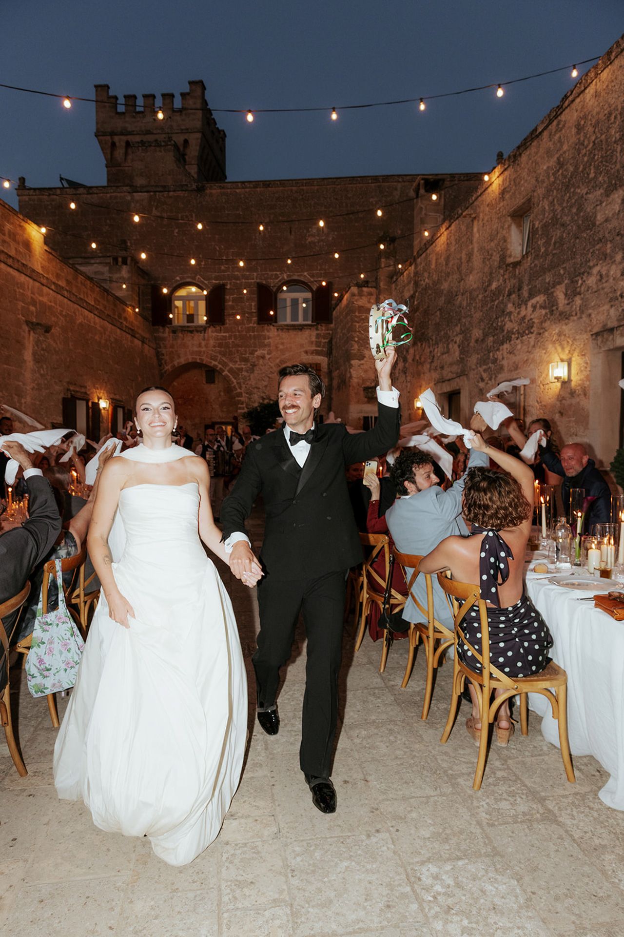 A wedding reception in a rustic stone venue with string lights overhead and guests dancing on a sandy floor.