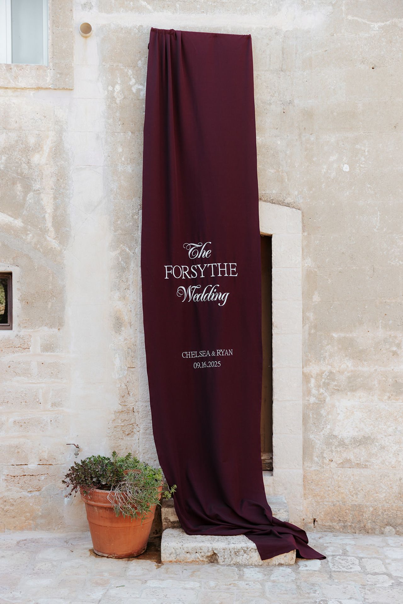 Burgundy wedding backdrop drapes against a textured stone wall with a decorative Forsythe wedding sign and potted plant.
