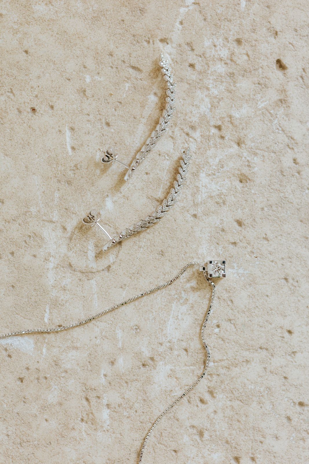 Delicate silver jewelry pieces including earrings and necklace with crystal details arranged on a beige textured surface.