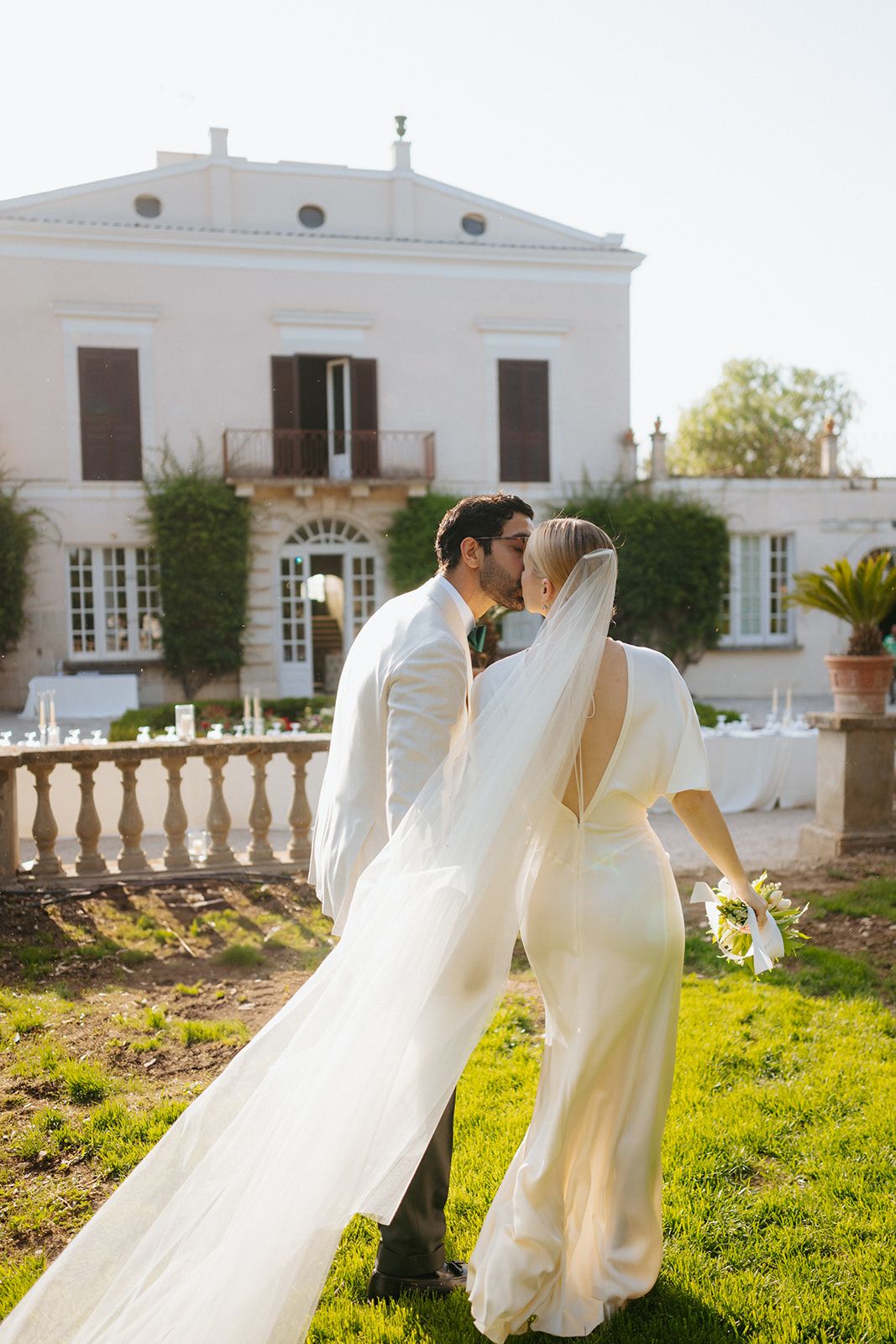 Elegant white villa provides romantic backdrop for intimate wedding moment in sunlit garden.