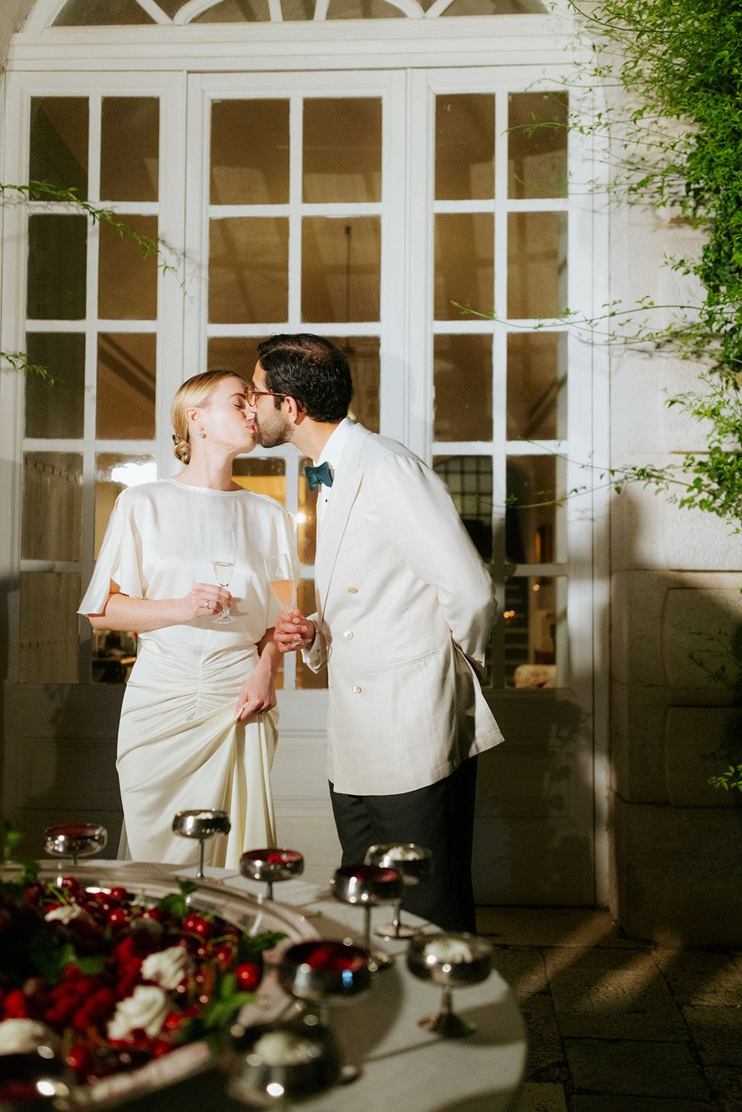 A romantic wedding moment with newlyweds sharing an intimate kiss by a glass-paned doorway at sunset.
