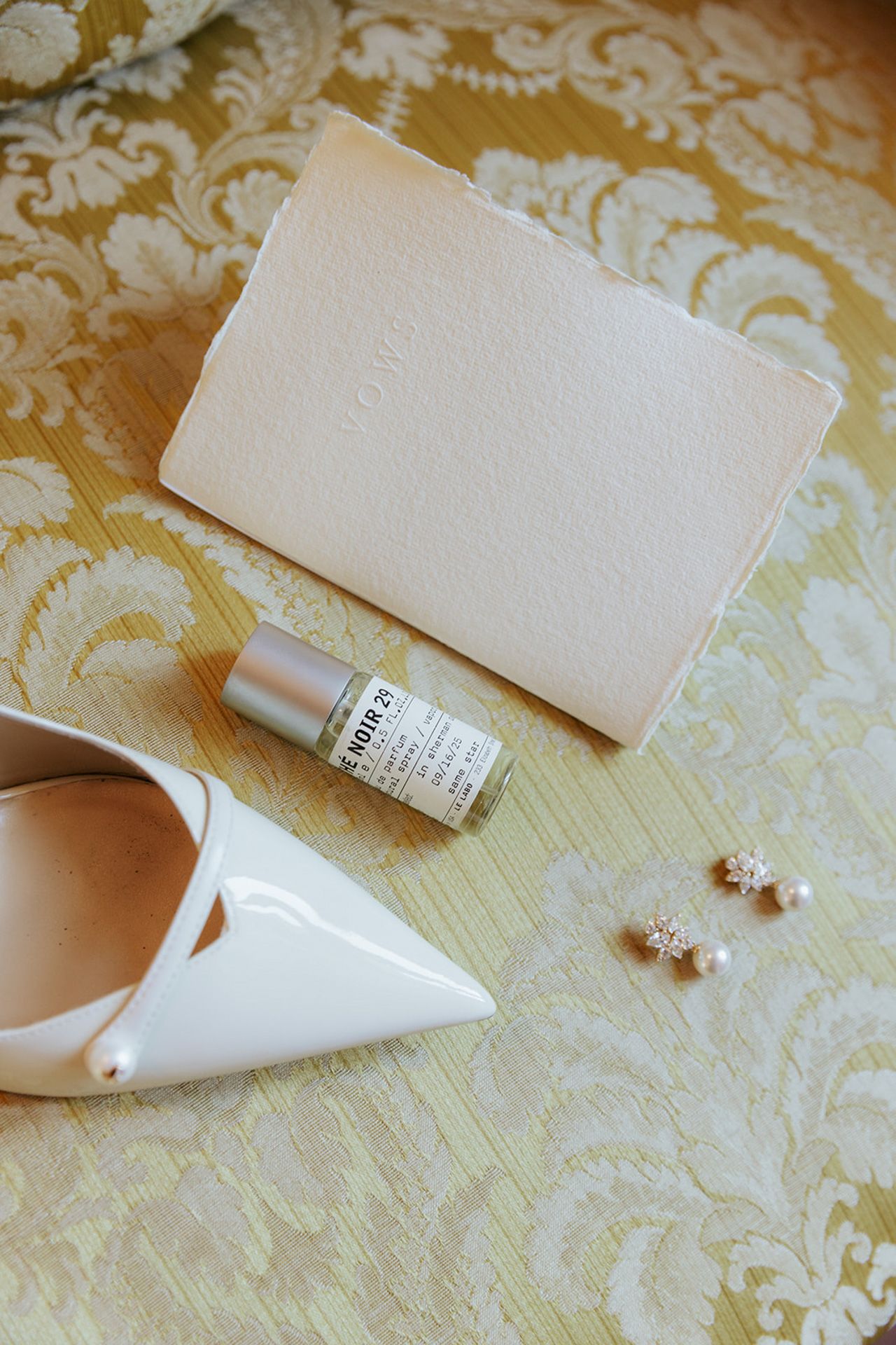 Wedding accessories including white peep-toe shoes, small perfume bottle and earrings on a damask patterned background.