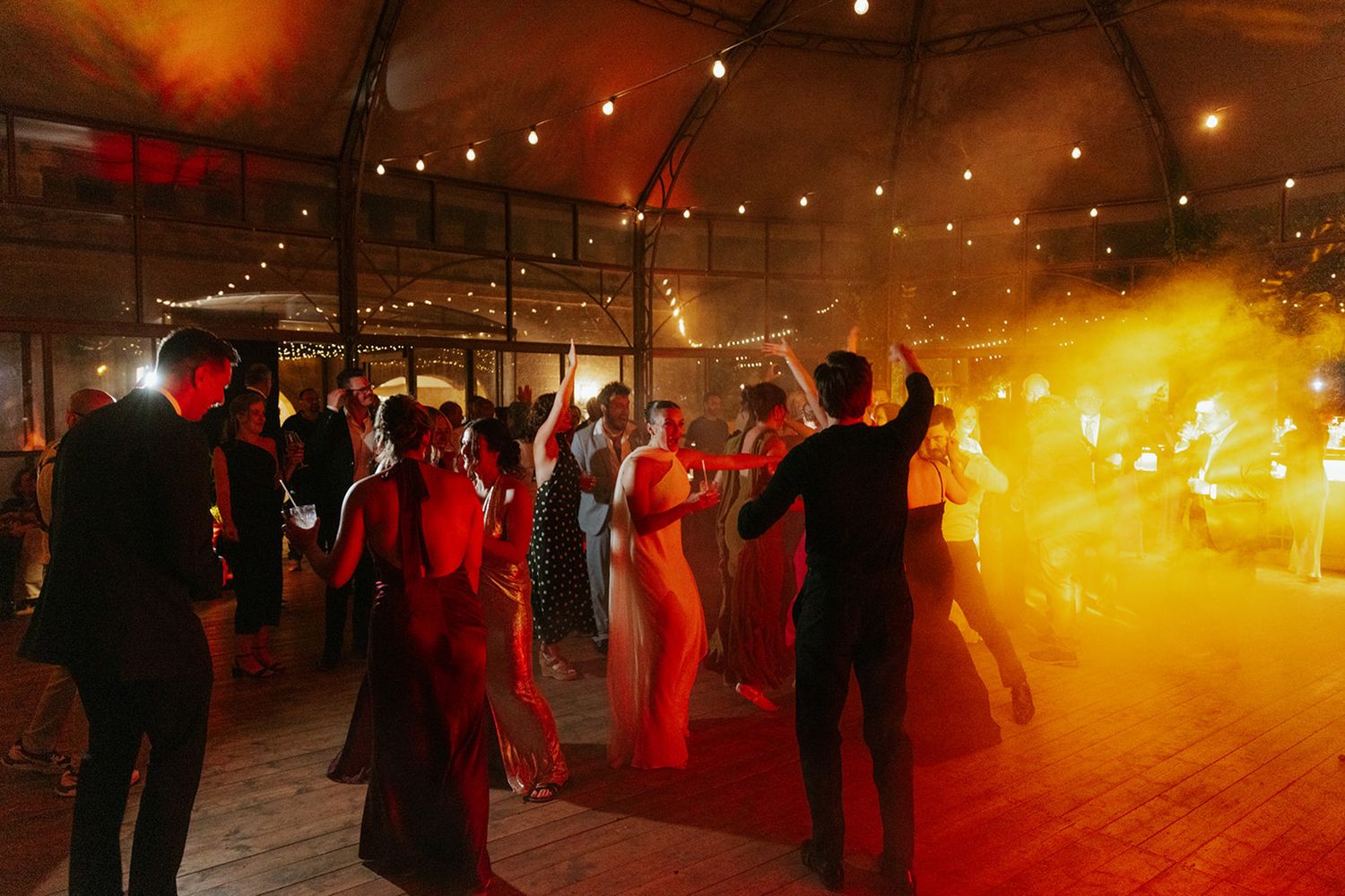 Wedding guests dance on a dimly lit dance floor illuminated by warm golden lights and smoke effects at an indoor reception.