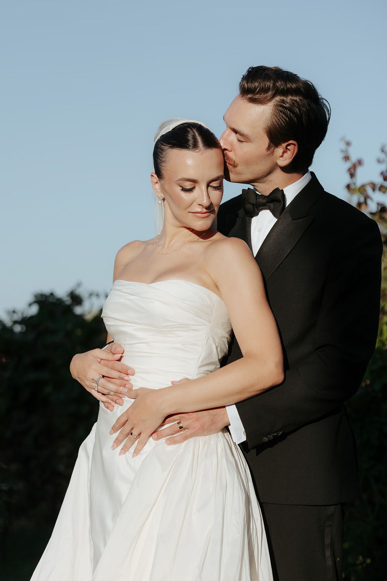 A tender moment between newlyweds as they embrace outdoors against a blue sky backdrop.