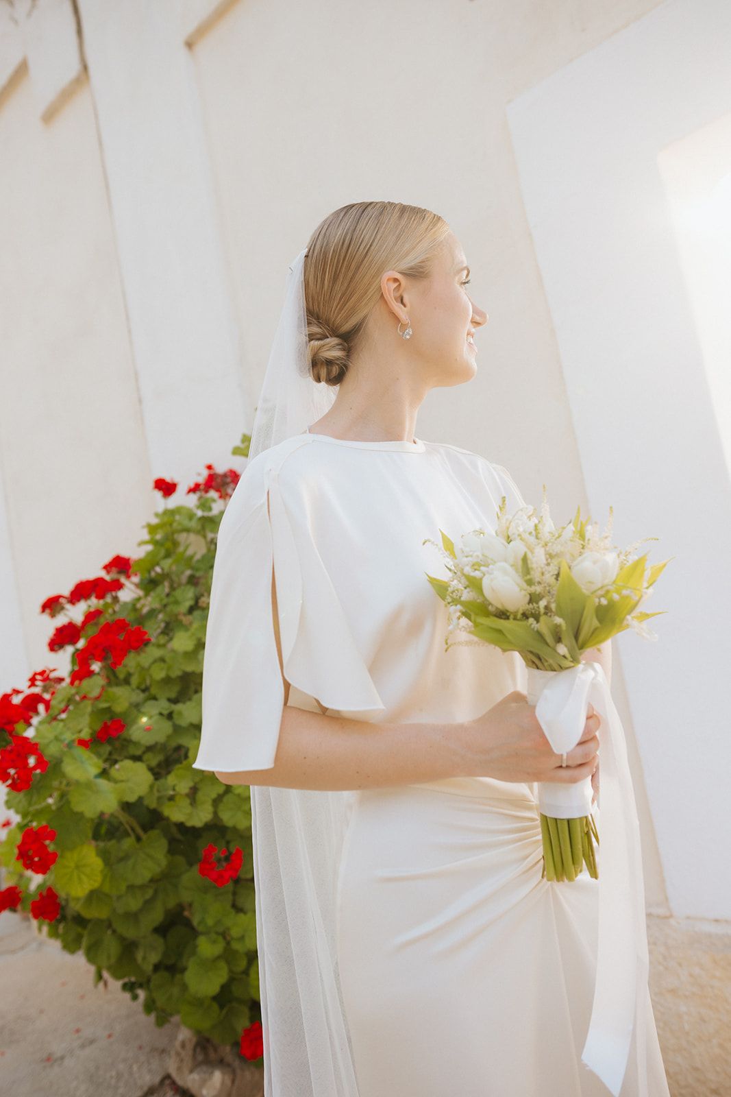 Elegant bride in white dress holds yellow lily bouquet while standing beside vibrant red geraniums in a sunlit garden.