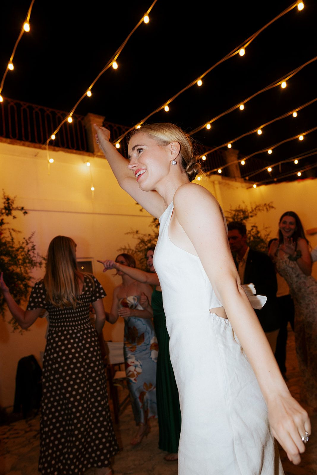 A festive outdoor party with string lights overhead and people dancing under the warm evening lighting.