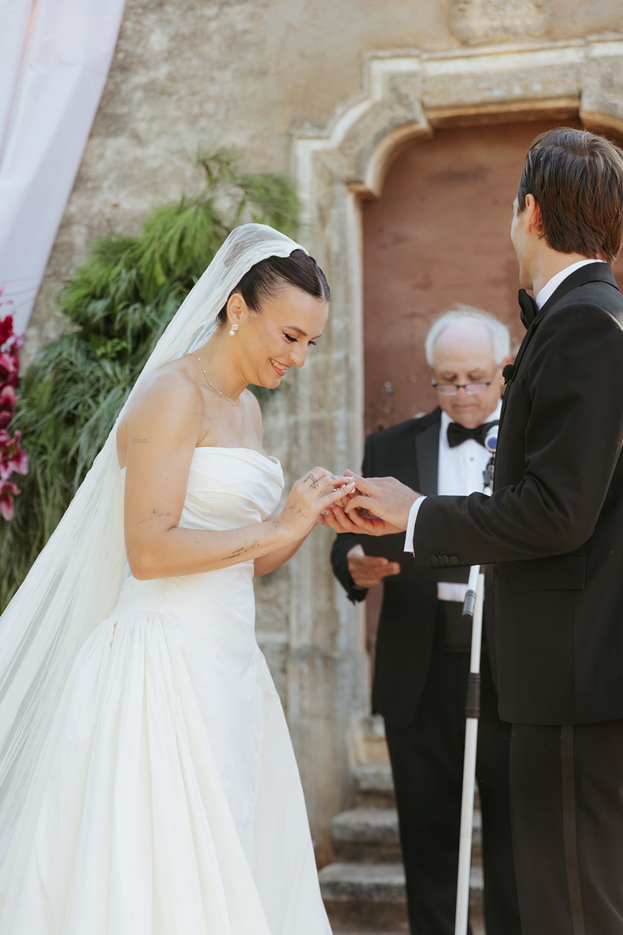 A wedding ceremony takes place outdoors with white draping and greenery decorations in the background.