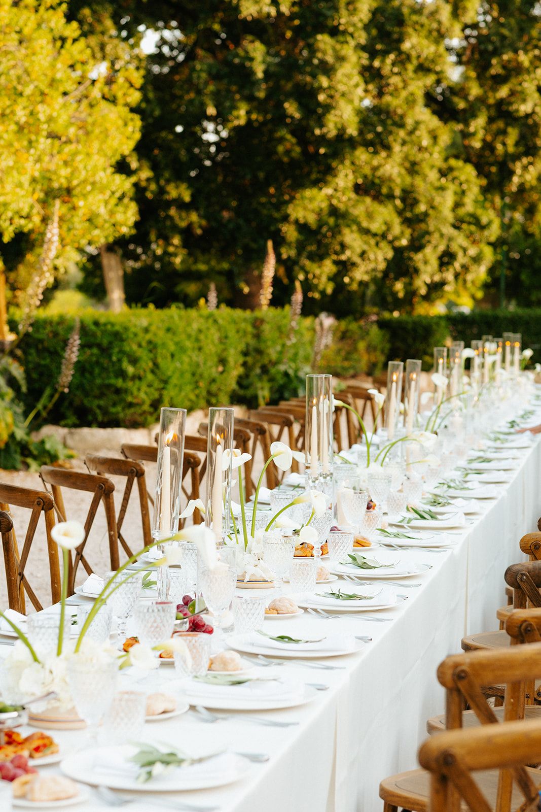 Elegant long banquet table set with white linens and rustic wooden chairs under golden sunlit trees for outdoor dining.