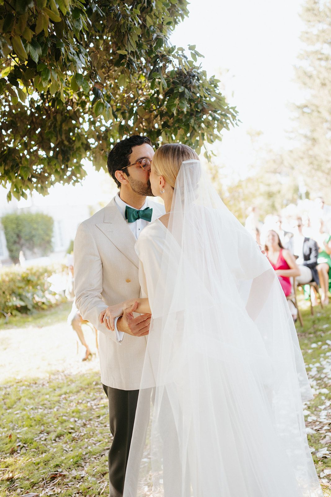 Two people in wedding attire share an intimate moment under a tree during an outdoor ceremony on a sunny day.