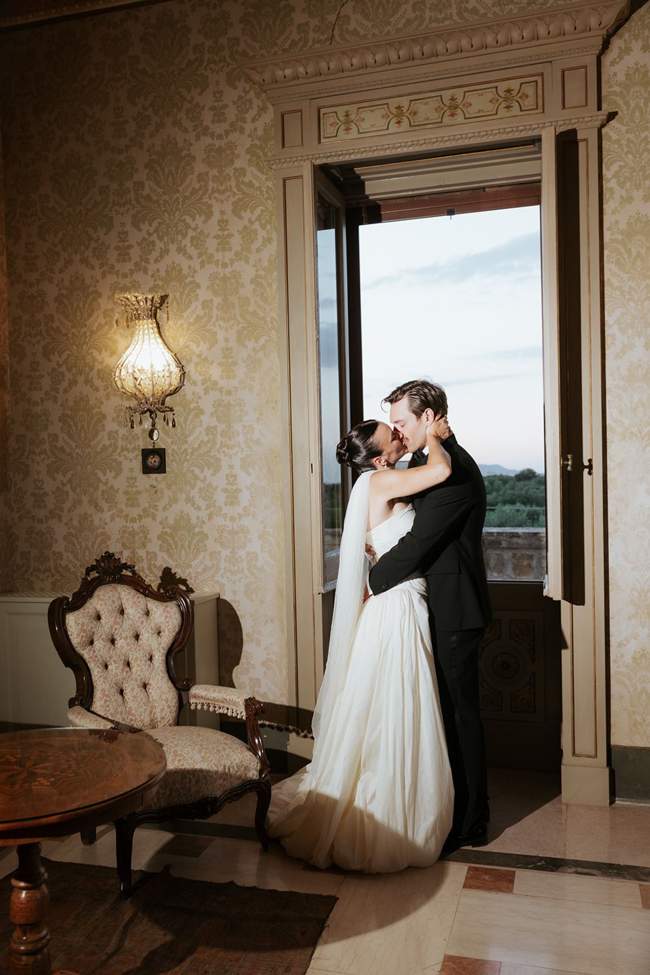 A romantic wedding couple embraces in a vintage room with ornate wallpaper and elegant antique furniture.