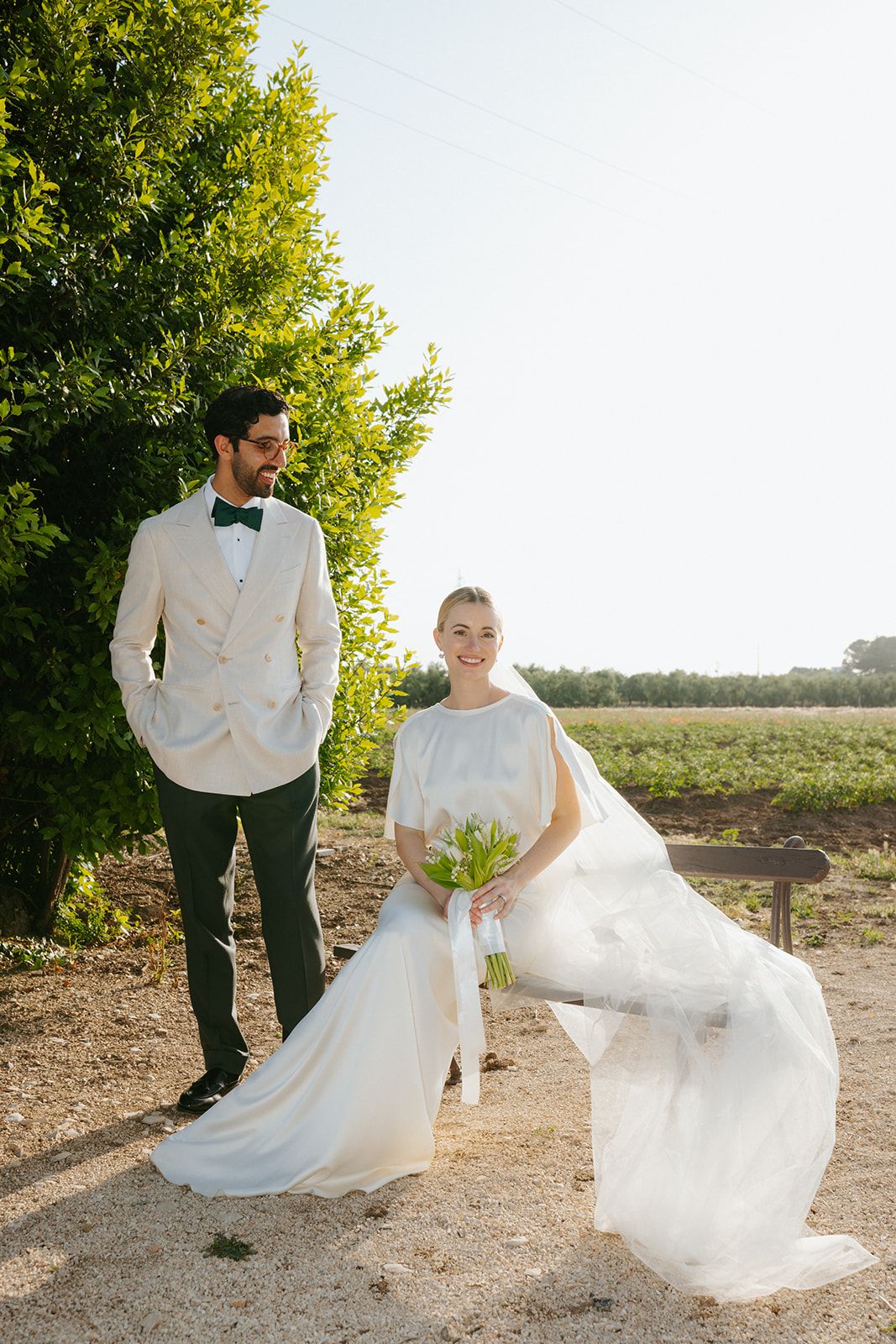 Wedding couple in white formal attire pose on a dirt path with green trees and sunlit field in background.