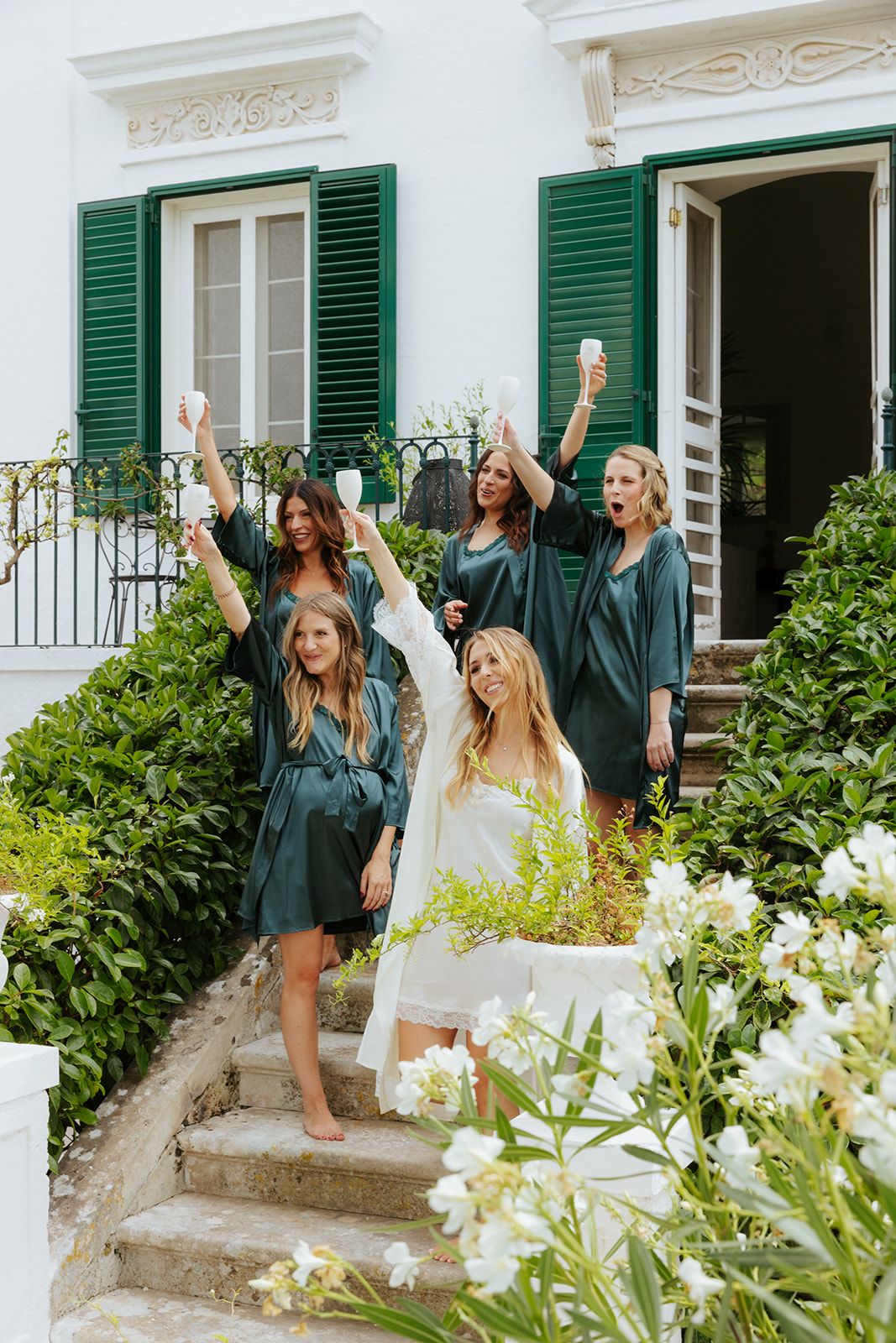 Graduates in dark green robes celebrate with raised arms on white building steps surrounded by flowering bushes.