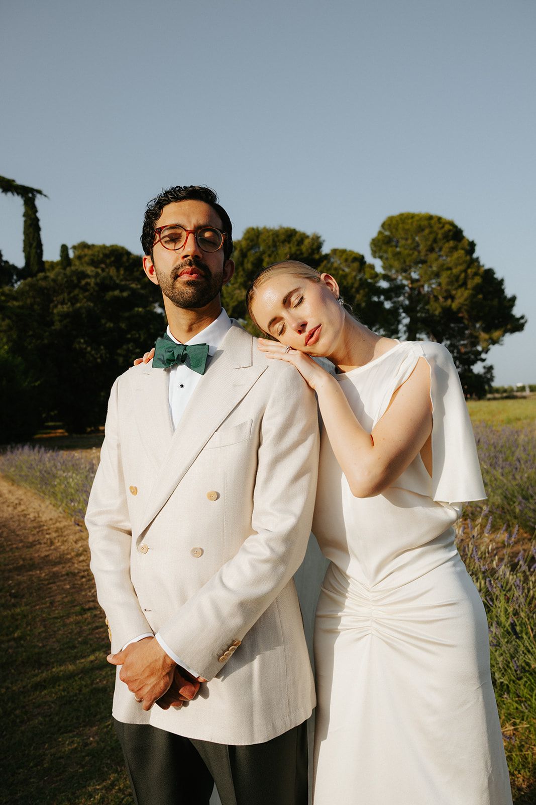 A couple poses in white formal attire against a scenic outdoor backdrop with trees and fields.