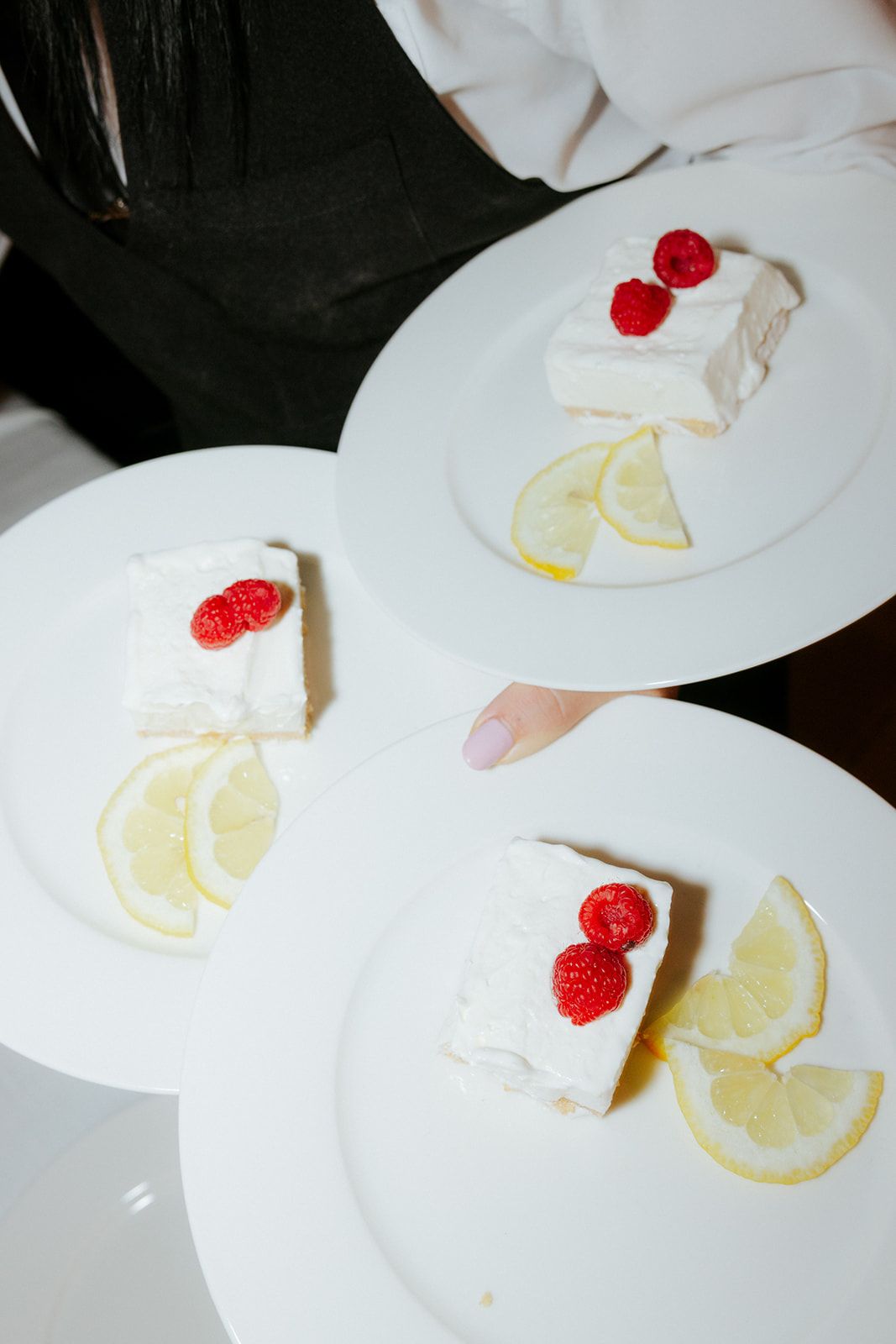 Three white plates with slices of cheesecake topped with fresh raspberries and lemon wedges garnish on a dark surface.
