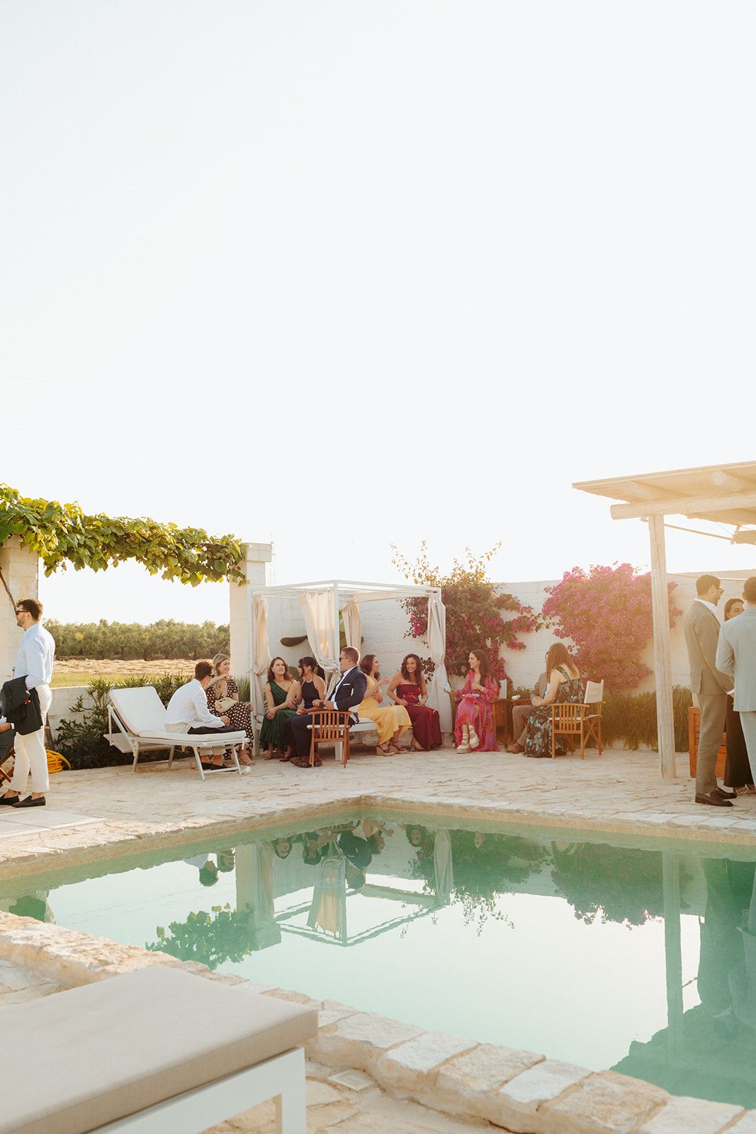 Sunny poolside gathering at a Mediterranean villa with people enjoying leisure time near a turquoise swimming pool at sunset.
