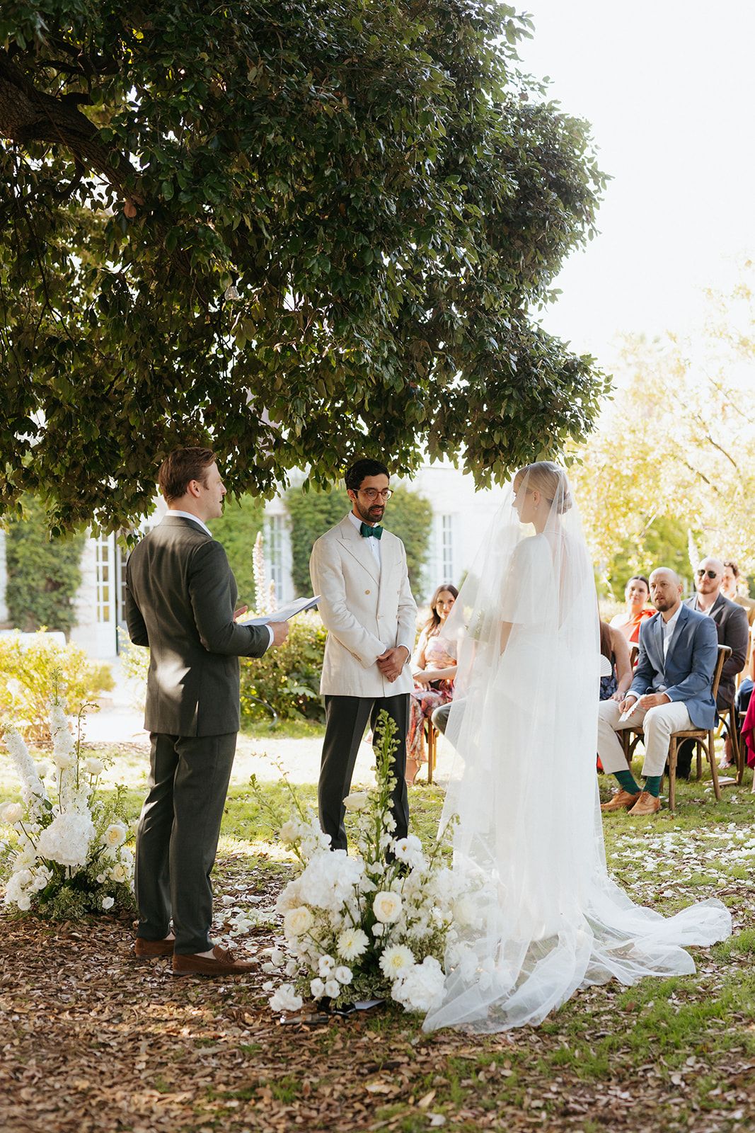 Outdoor garden wedding ceremony with flowers and guests seated under trees on a sunny day.