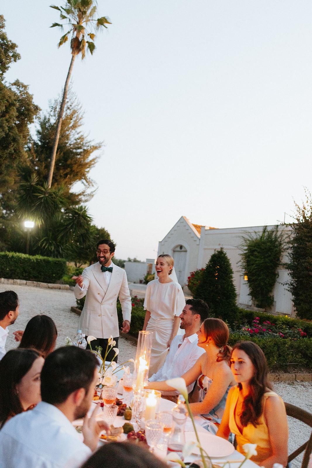 Guests enjoying an elegant outdoor dinner party at sunset with white decor and candlelit tables.