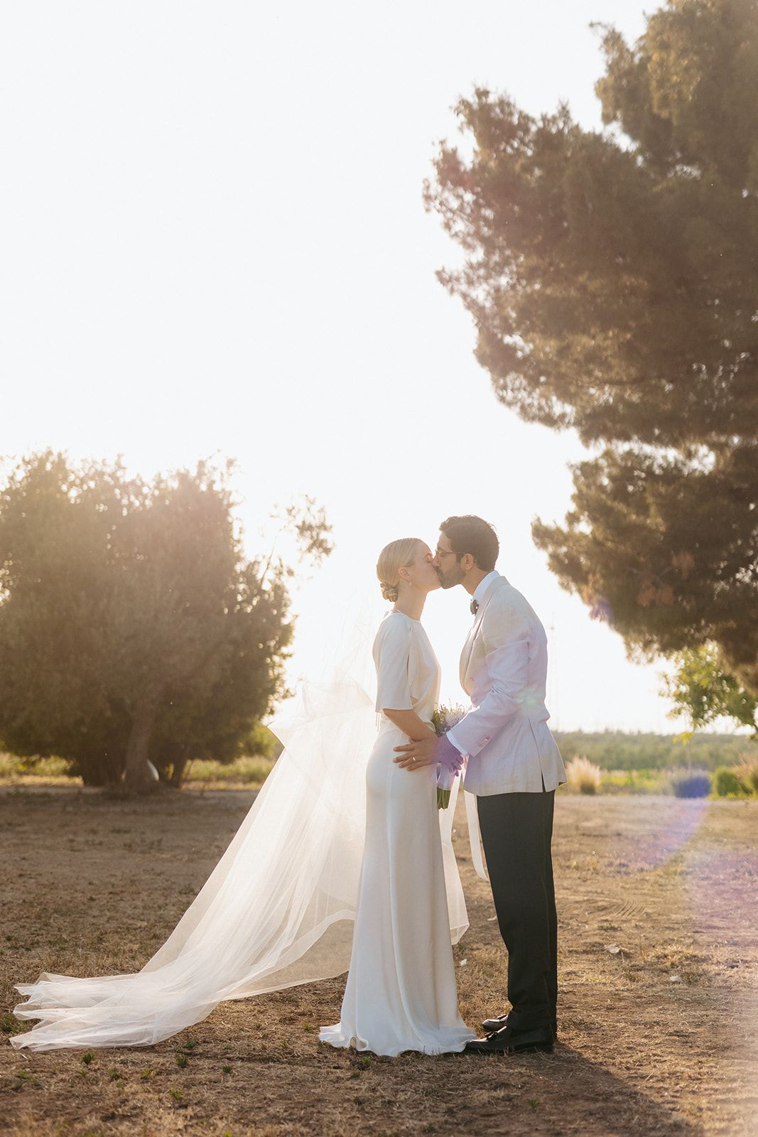 Romantic sunset wedding photo of a couple kissing in an outdoor garden setting with soft golden light filtering through trees.
