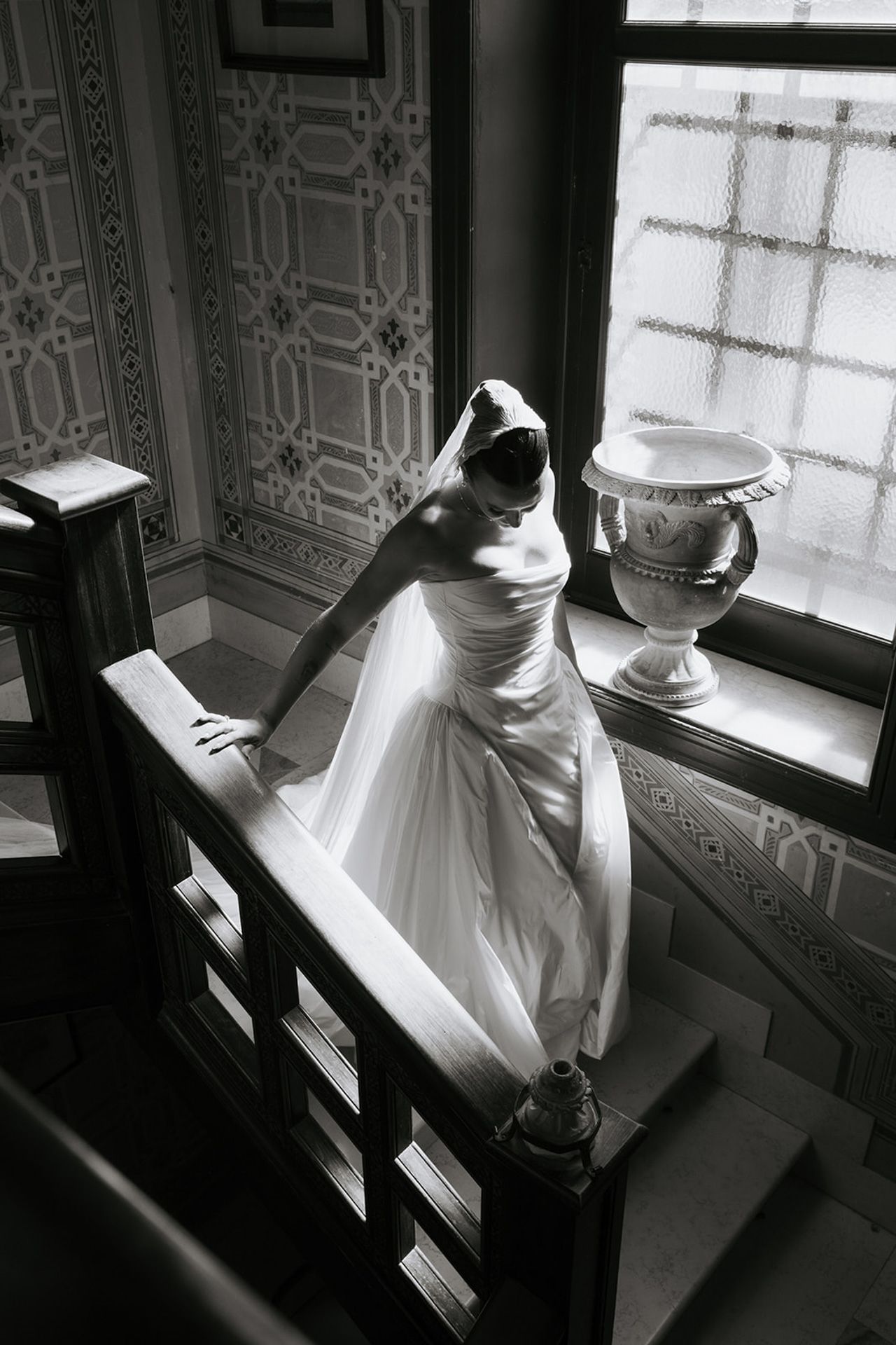 A black and white photograph of a flowing white dress on stairs near an ornate window with a decorative urn.