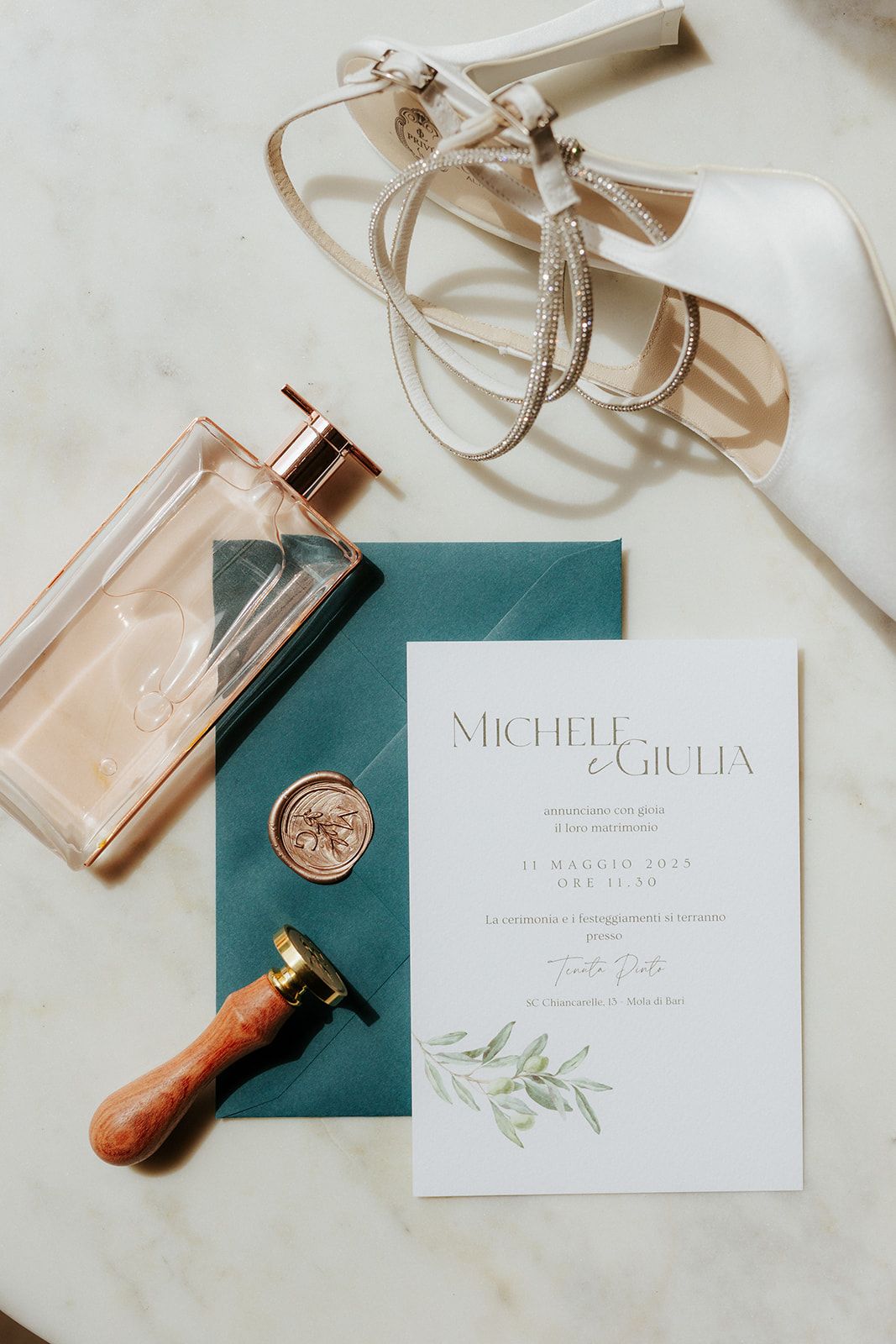 Elegant wedding invitation with wax seal on teal paper alongside perfume bottle, white heels and wooden stamp on marble surface.