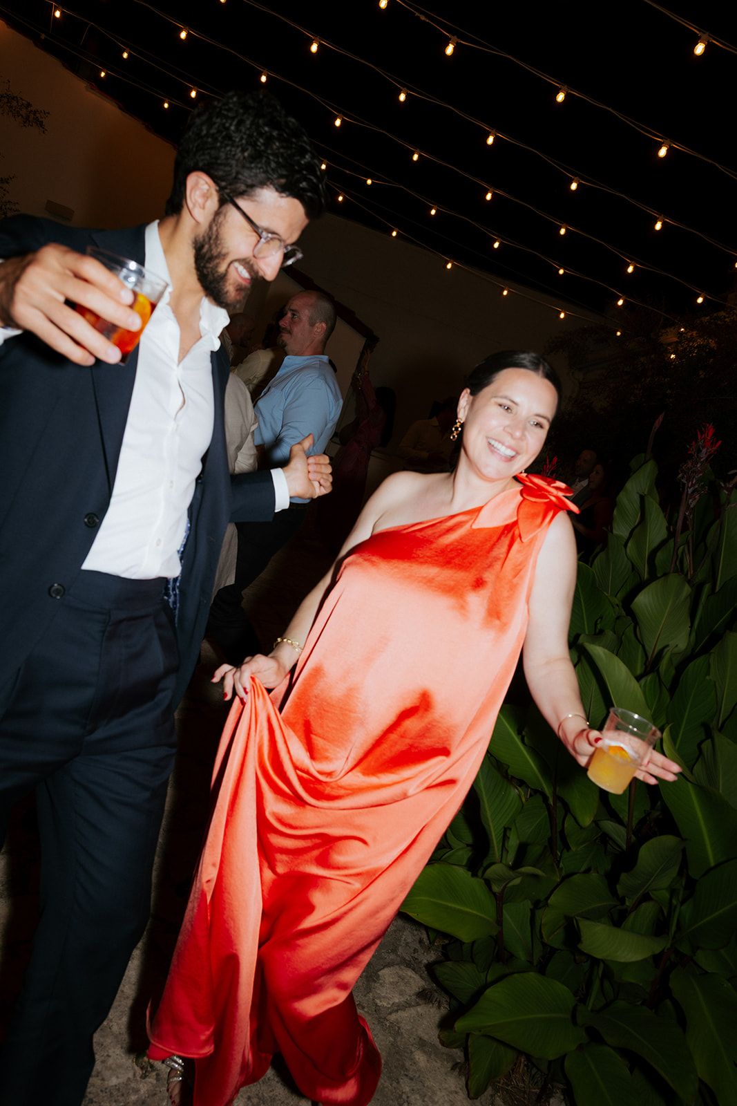 People dancing at a party under string lights with someone wearing a vibrant orange satin dress.