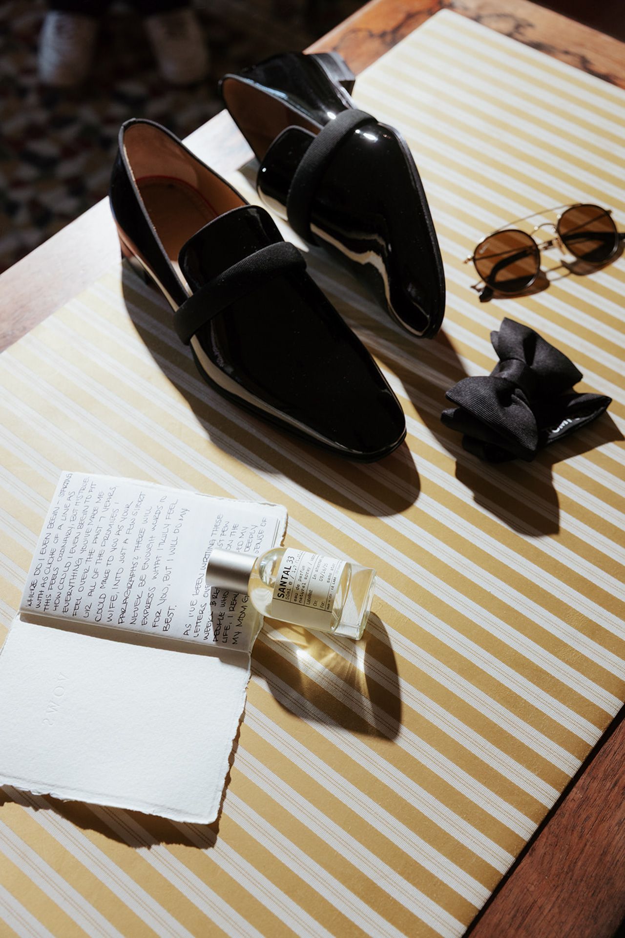 Black leather loafers and a perfume bottle arranged on a striped yellow and white surface in natural lighting.