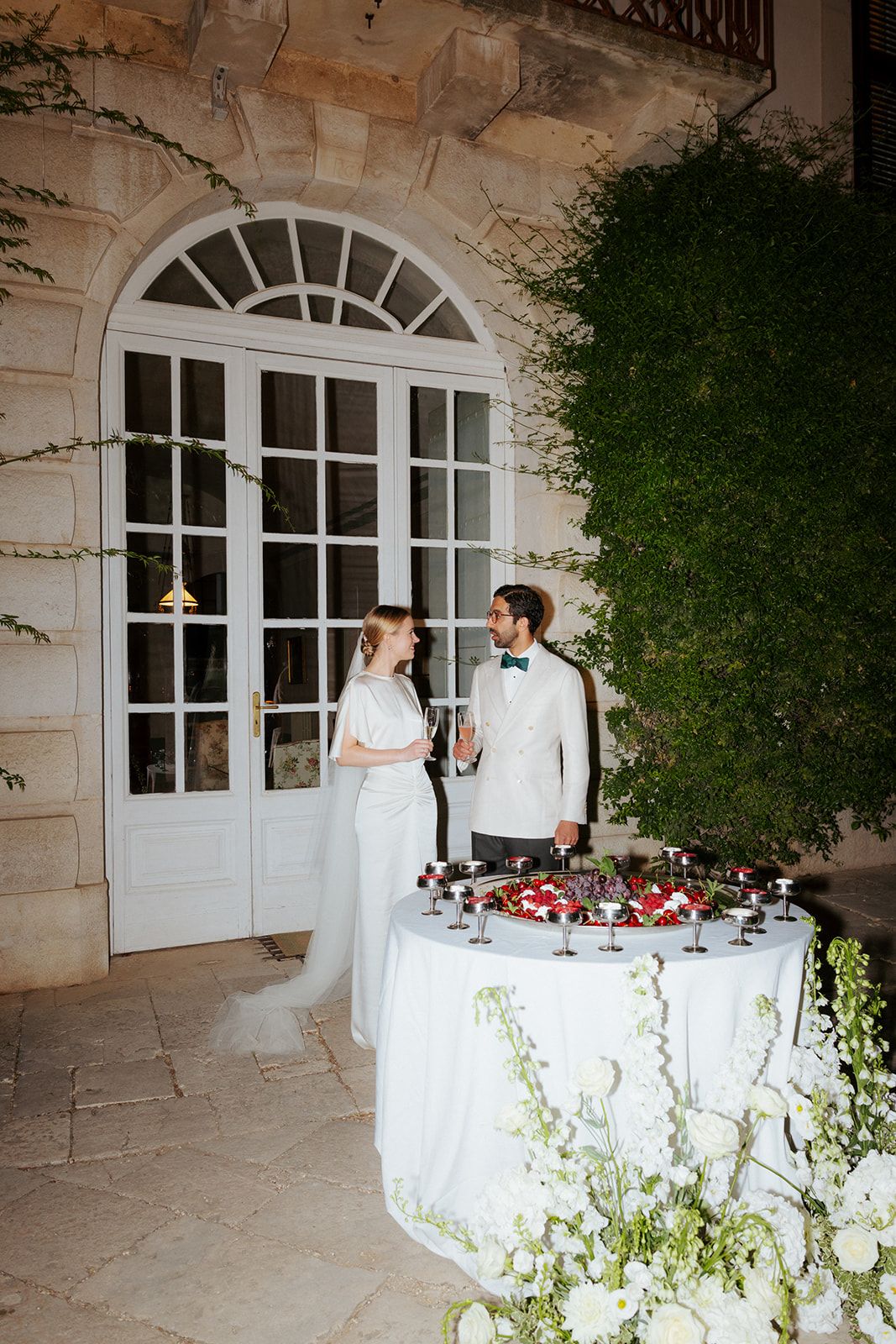 An elegant reception table draped in white linens with floral arrangements sits outside a stone building with arched windows.