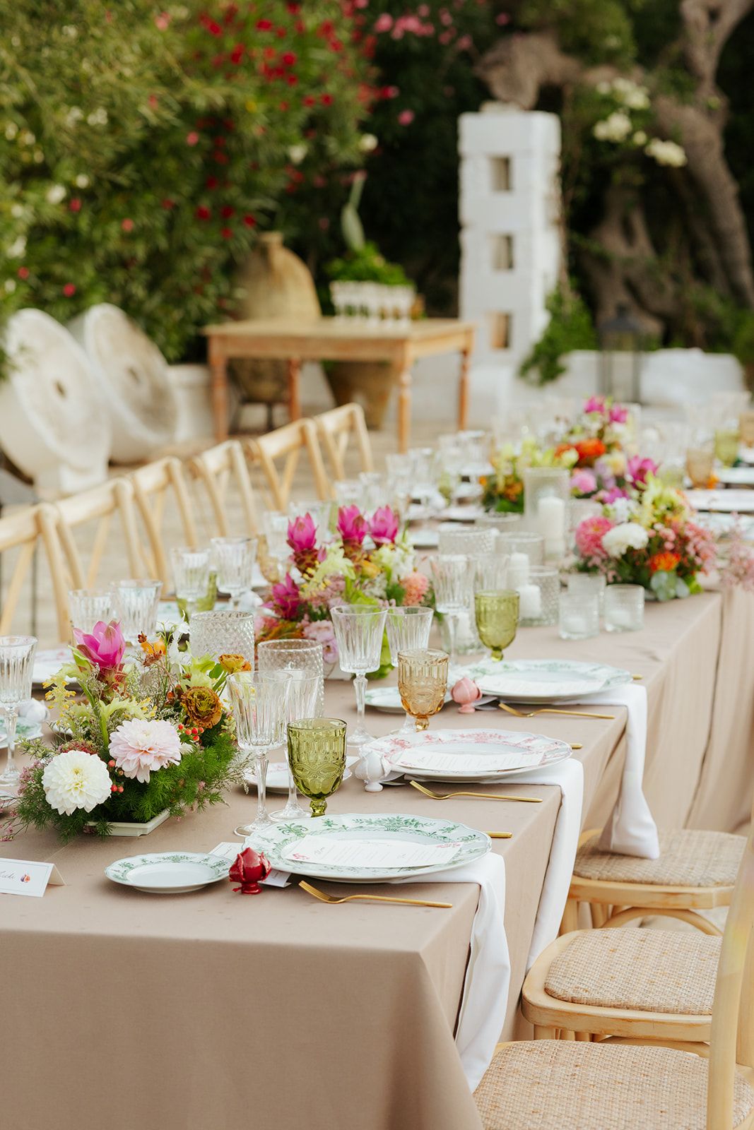 Elegant outdoor wedding reception table settings with beige linens, wooden chairs, and colorful floral centerpieces.