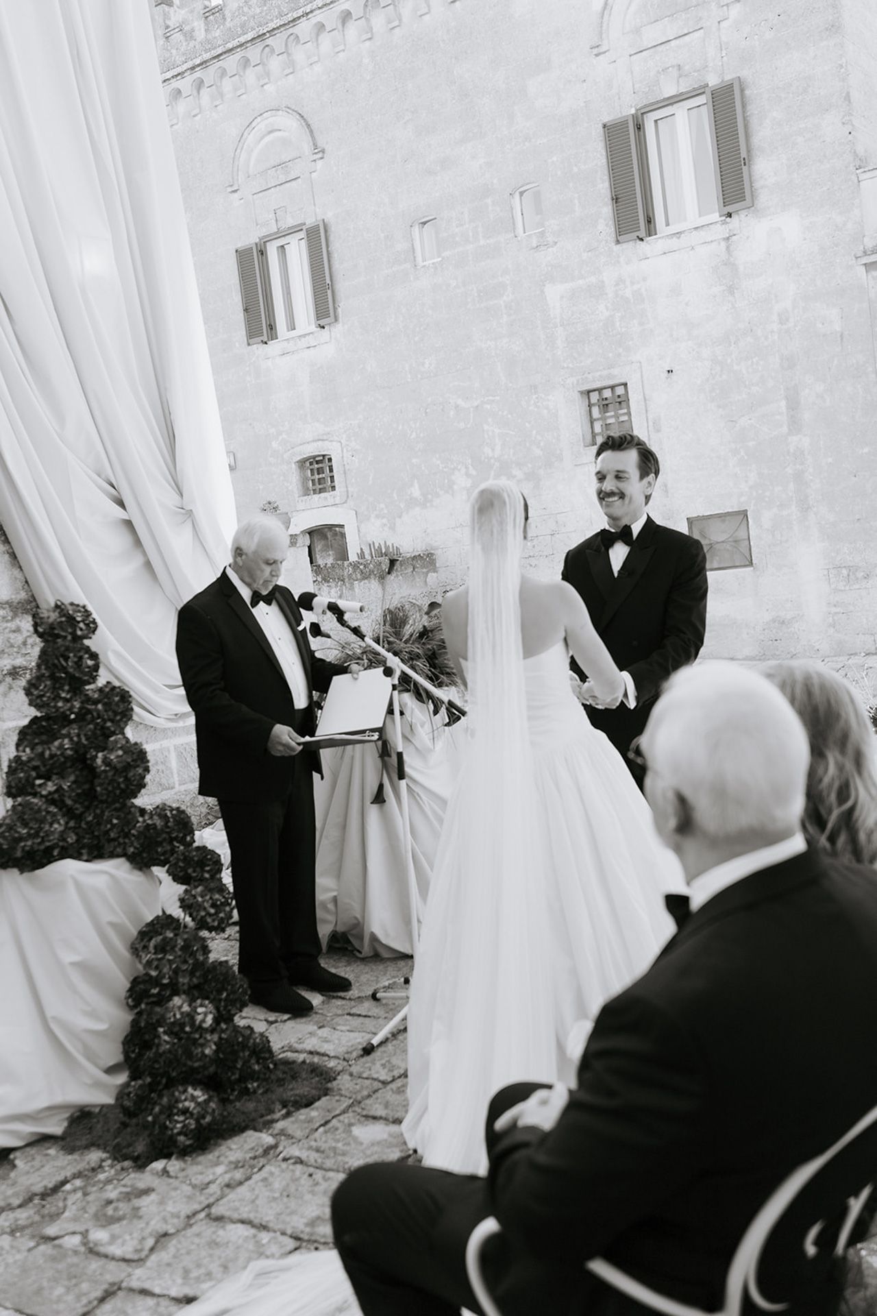 A series of black and white wedding photographs showing a couple and guests outside a stone building.