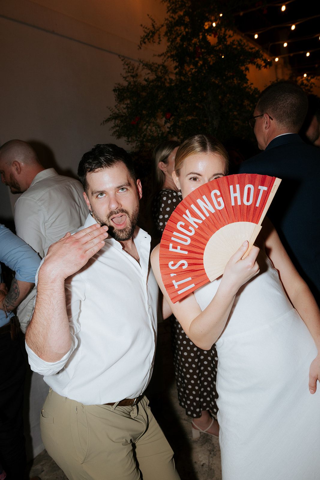 Party guests dance beneath string lights with some holding red fan props during an indoor celebration.