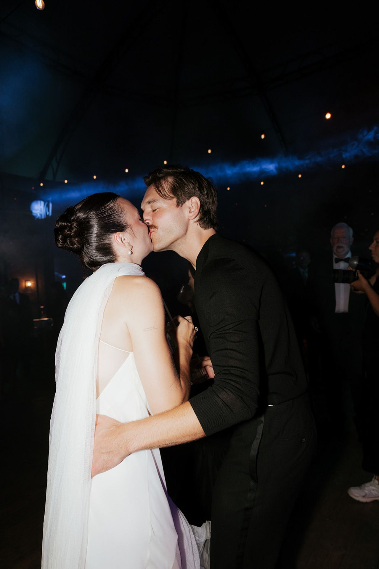 A romantic first dance moment between newlyweds sharing a kiss at their wedding reception with blue lighting in the background.