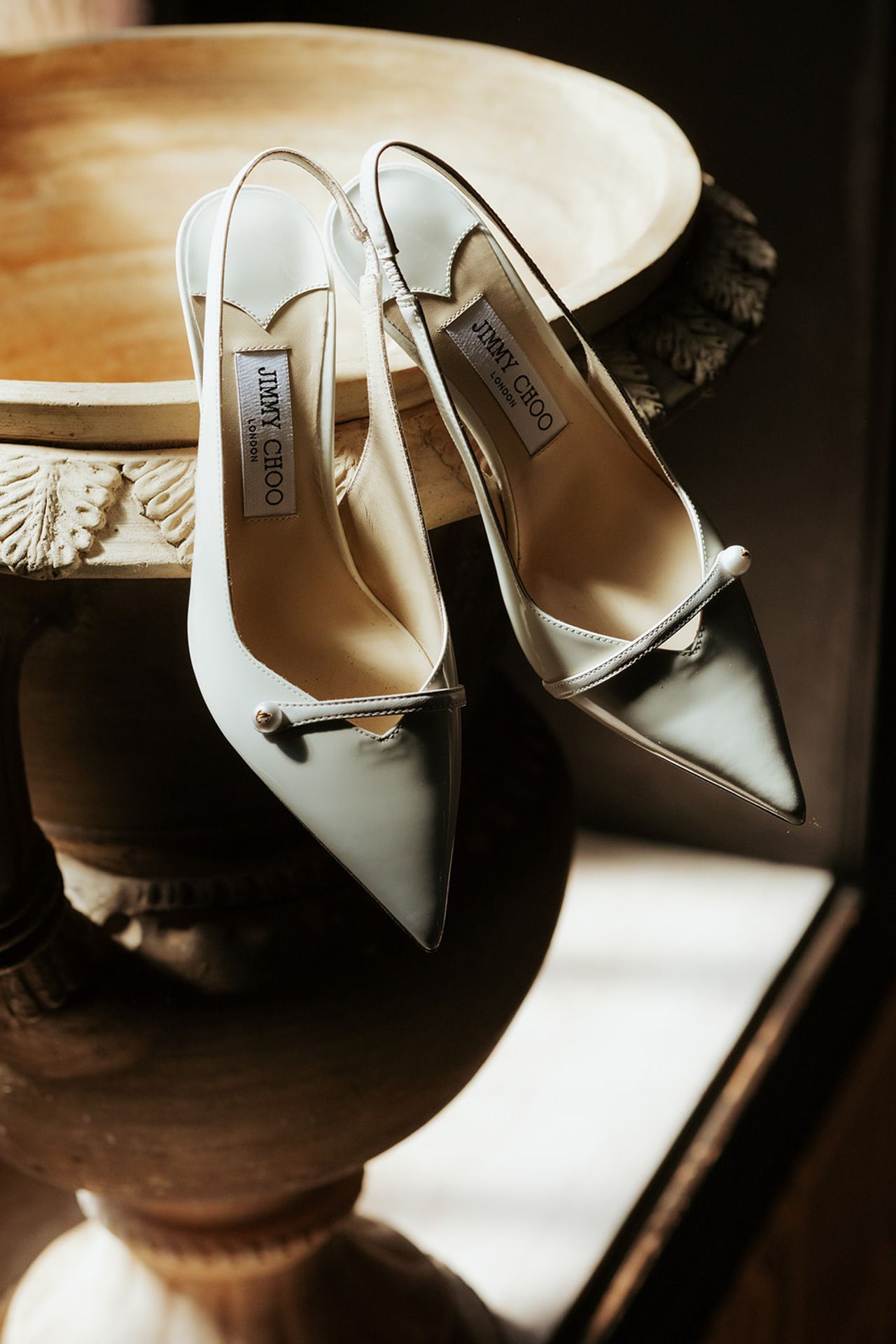 Elegant white Jimmy Choo slingback heels with pointed toes and bow details sit on a wooden surface in dramatic lighting.