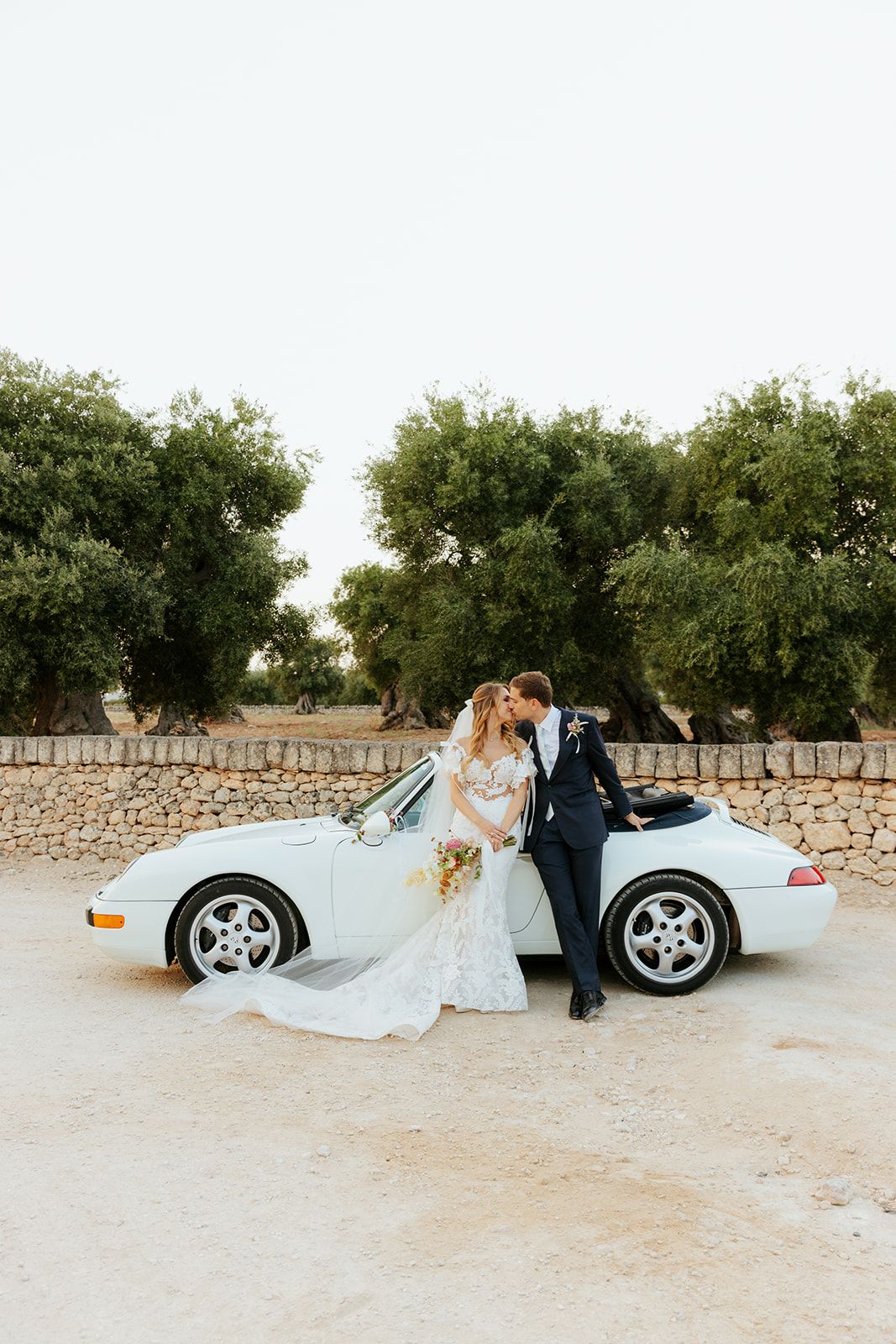 A wedding couple poses artistically near a vintage convertible car on a sandy road with olive trees in the background.