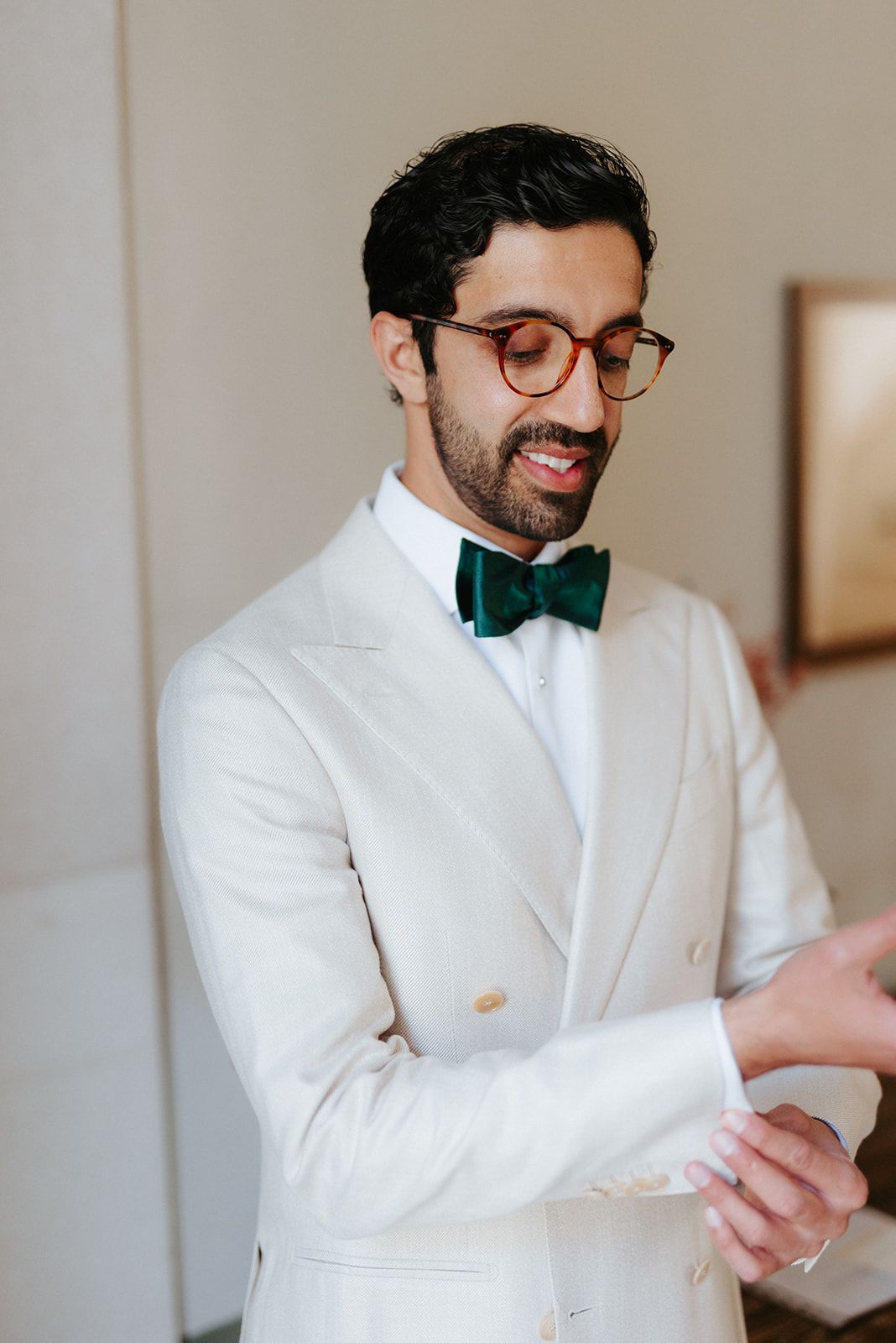 Groom in a crisp white double-breasted suit and emerald green bowtie adjusts his cuffs while smiling.