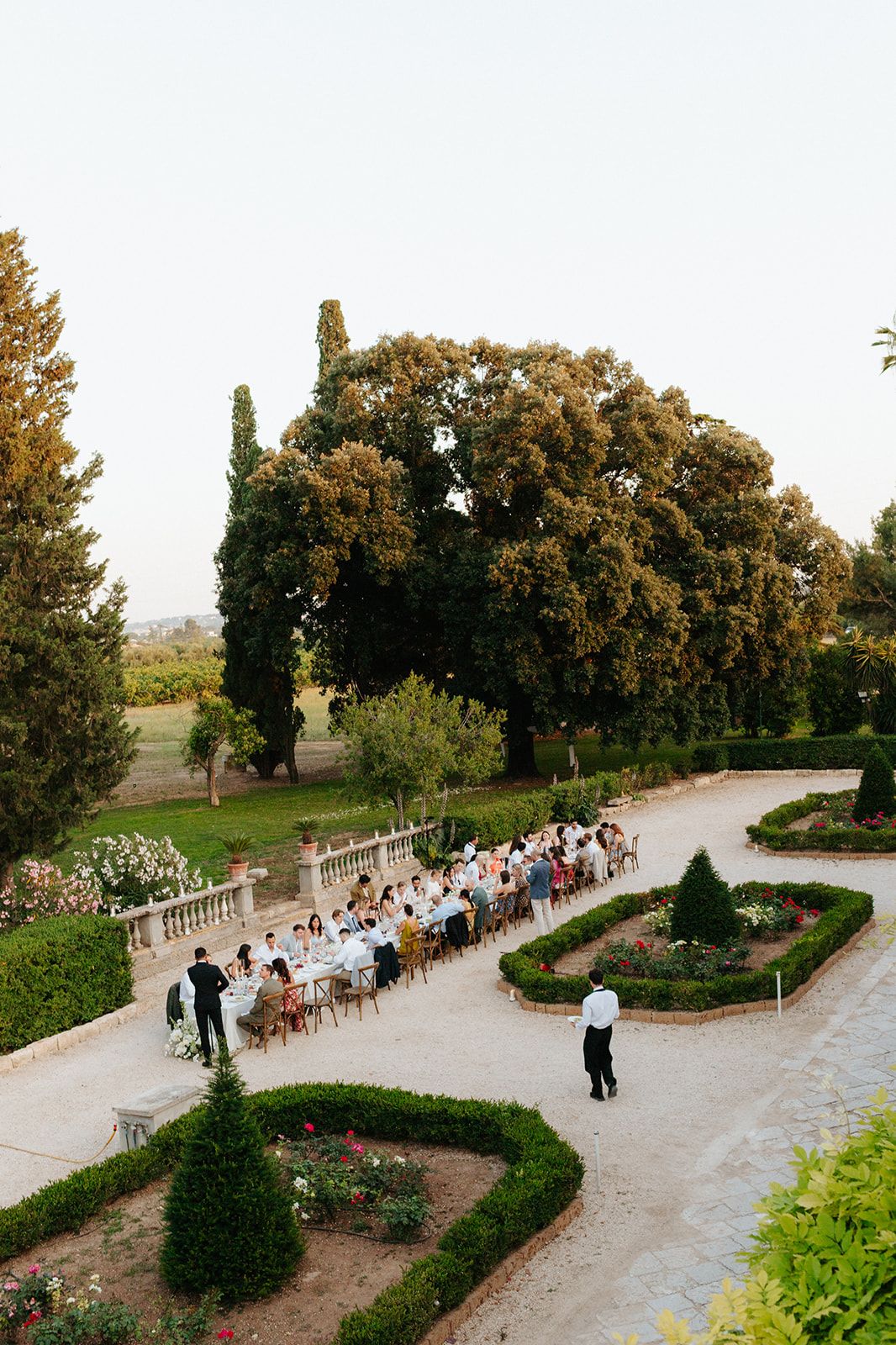Overhead view of an elegant outdoor wedding reception with long tables set up along a curved garden path surrounded by manicured hedges.