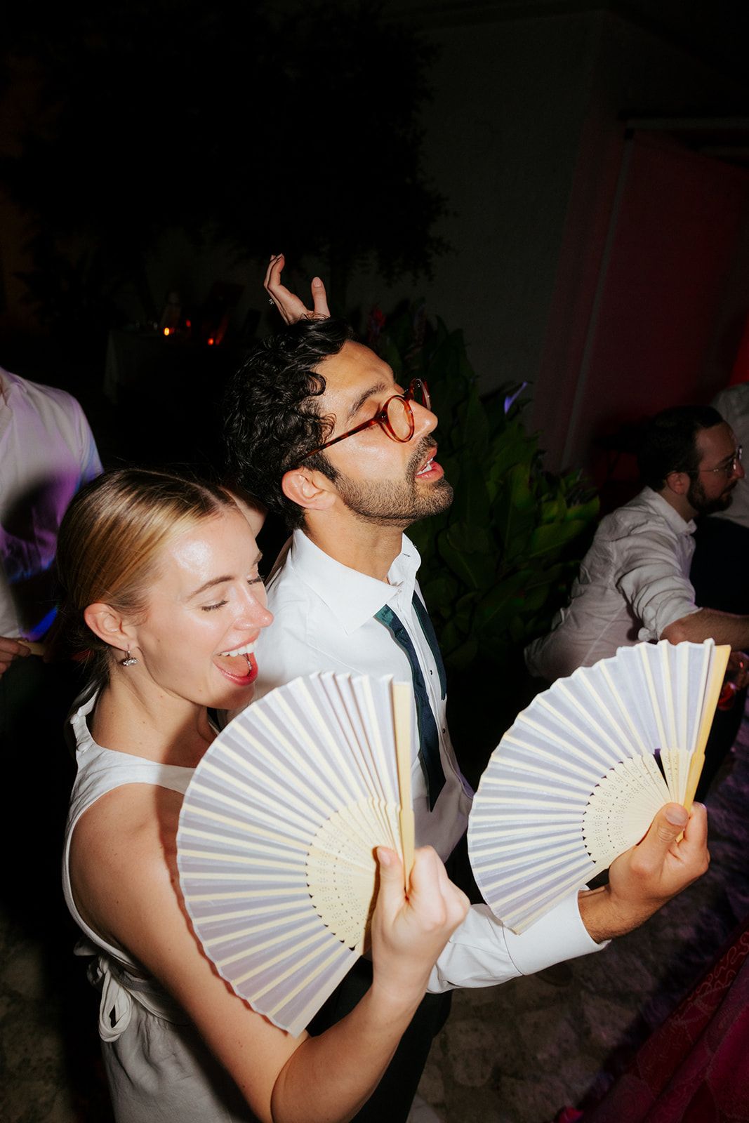 Two party guests in formal attire dance with white hand fans at a dark indoor celebration.