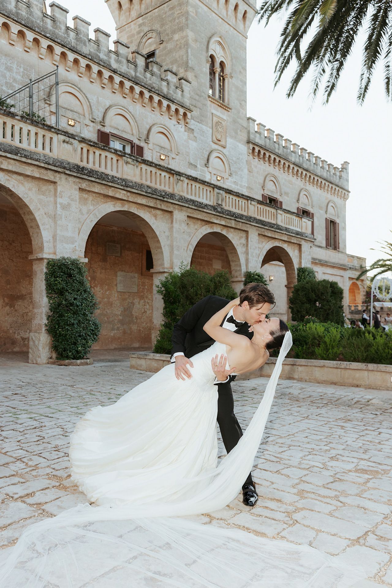 A romantic wedding photo at a Mediterranean castle with stone archways and palm trees.