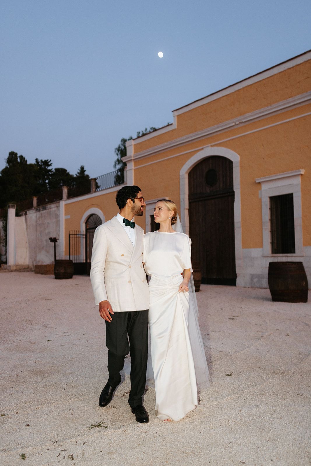 A romantic wedding couple in white attire poses in front of a beige Mediterranean-style building at dusk.