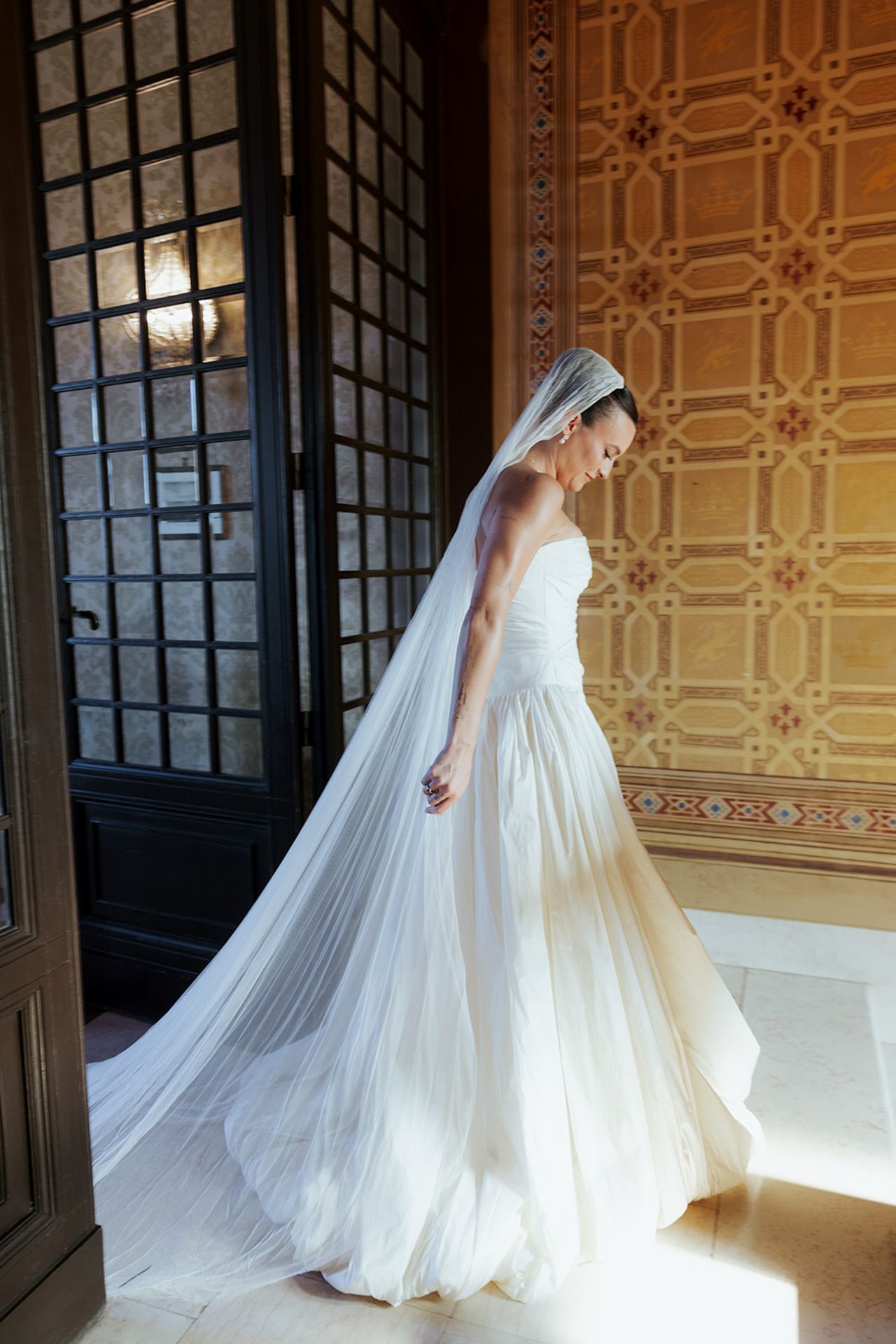A bride in a flowing white wedding gown and long veil stands in elegant hallway with ornate wallpaper and dark doorframe.
