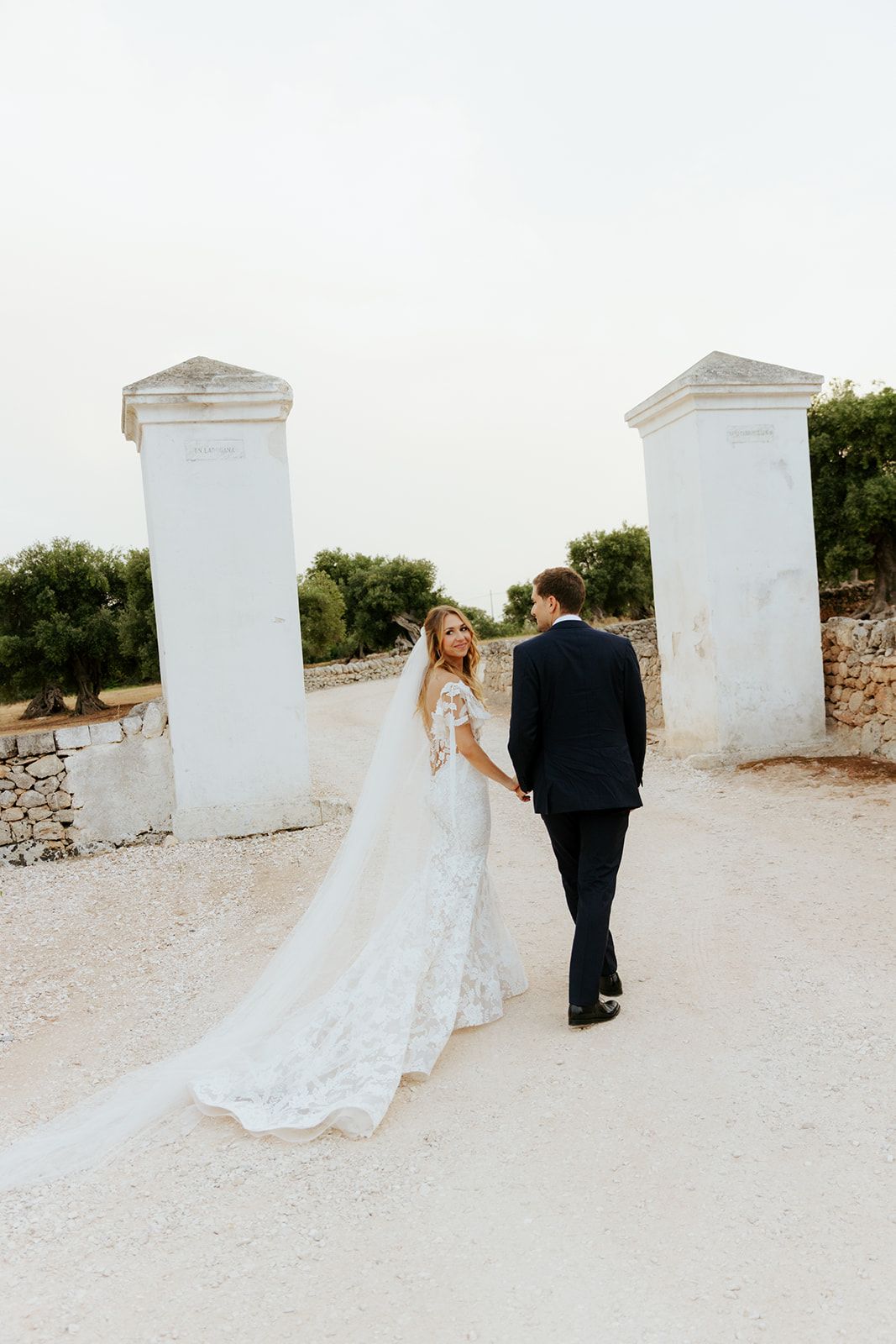 Wedding couple walks along white Mediterranean style walls on rustic gravel path.