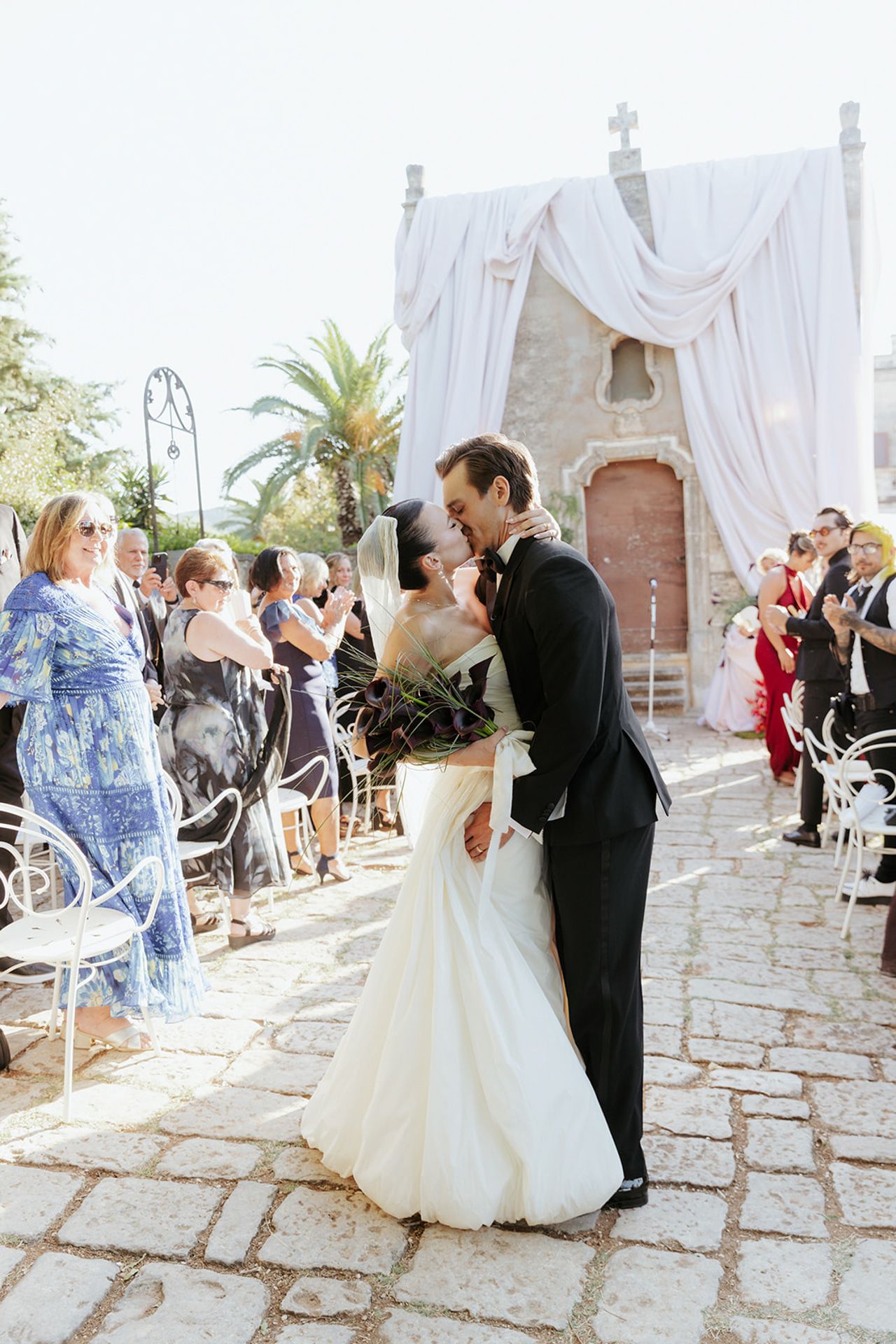 A romantic outdoor wedding ceremony with white draped decor, stone pathway and palm trees in the background.