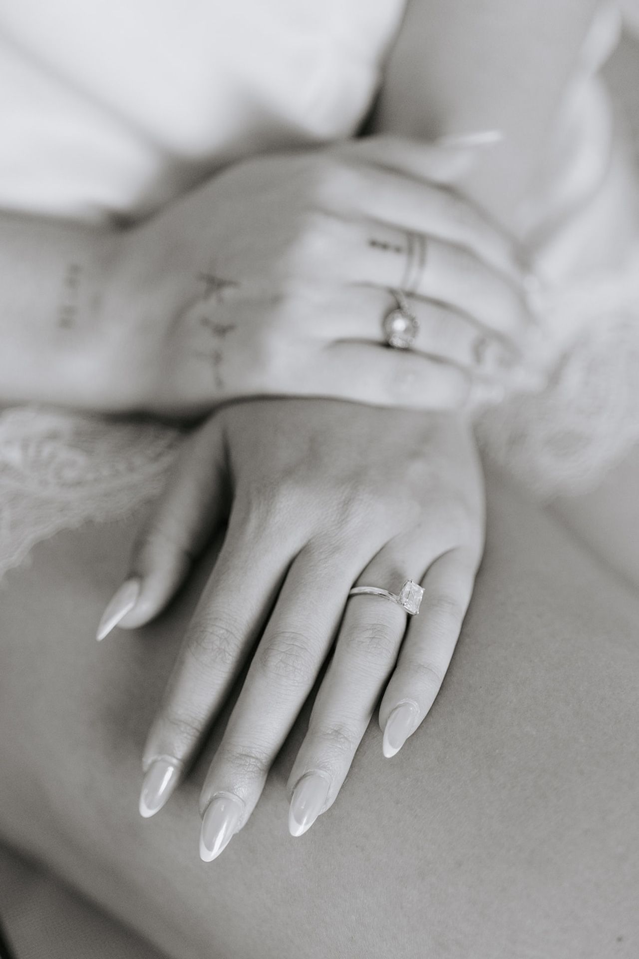 Black and white close-up photograph of hands with long manicured nails resting on a soft surface.