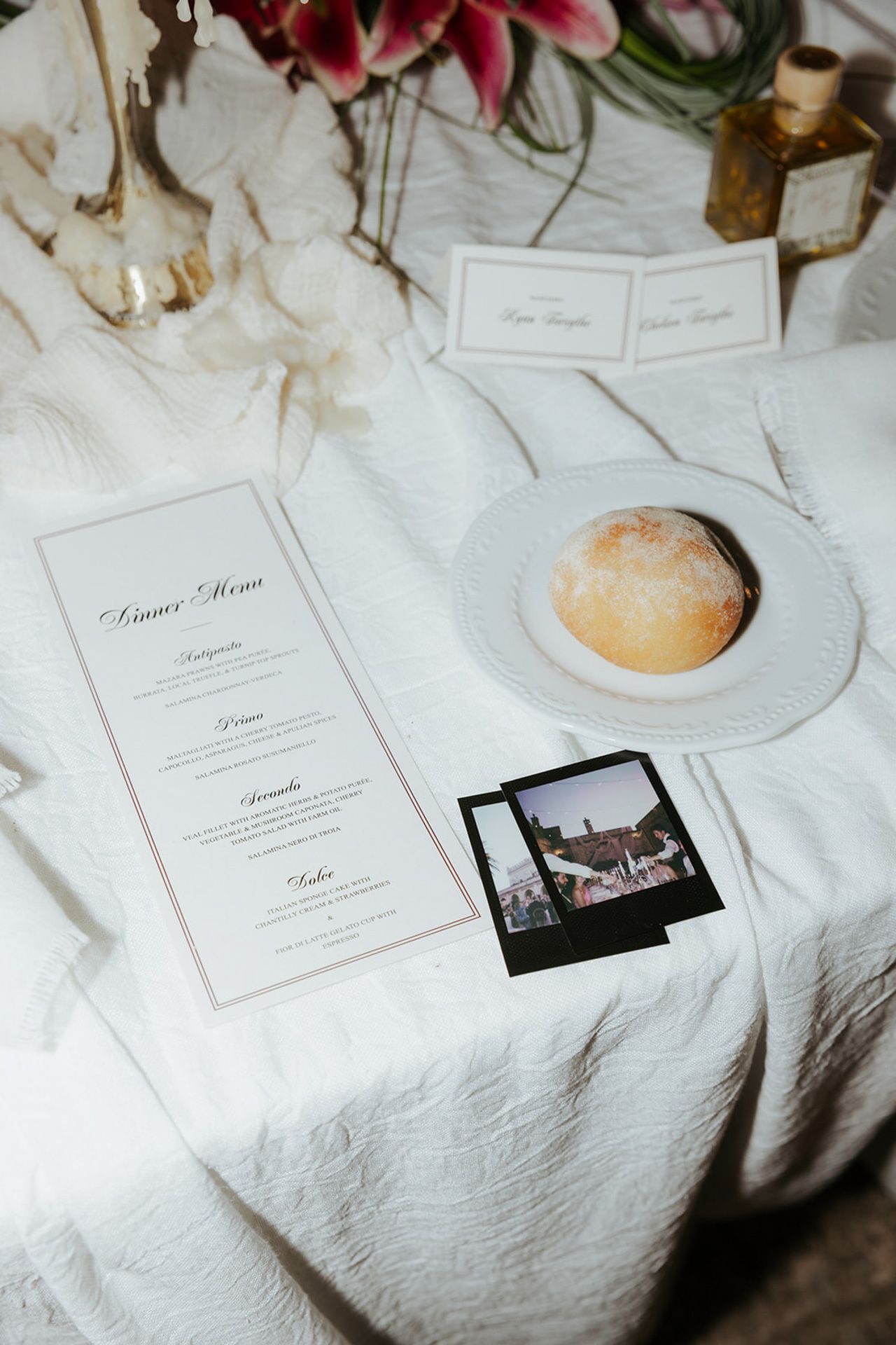 A dinner menu card lies on a white tablecloth beside a plate with bread and vintage polaroid photos.